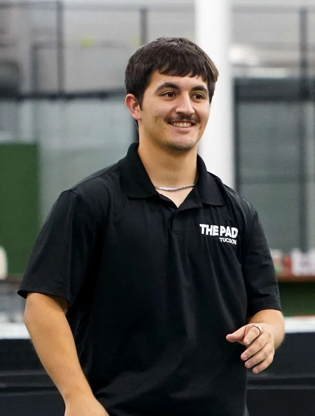 Person playing pickleball indoors, wearing a black hoodie, shorts, and a white cap. Focused on a yellow pickleball near their paddle on a green court. Coach Greg Vinueza