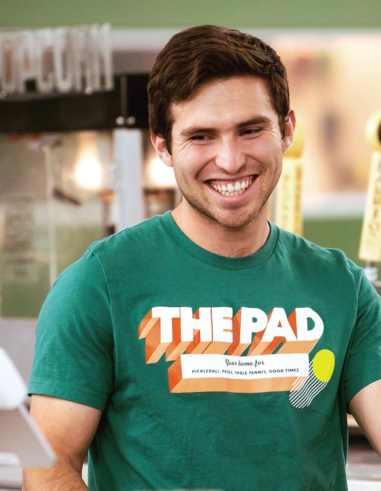 Person playing pickleball indoors, wearing a black hoodie, shorts, and a white cap. Focused on a yellow pickleball near their paddle on a green court. Coach Greg Vinueza