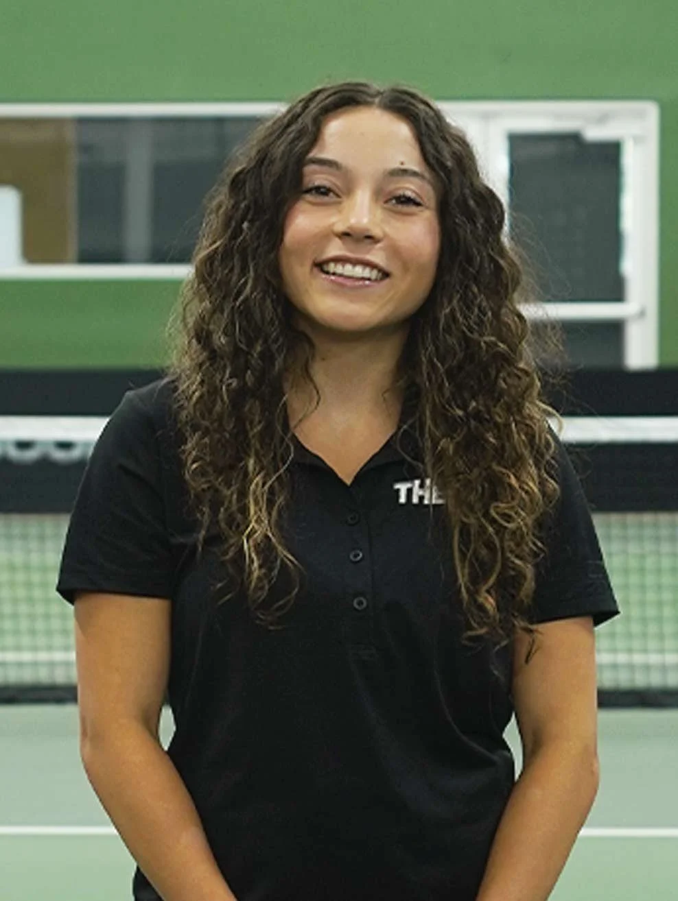A young woman playing pickleball indoors wearing a green shirt with "THE PAD TEAM" text. Coach Tyler Roop.