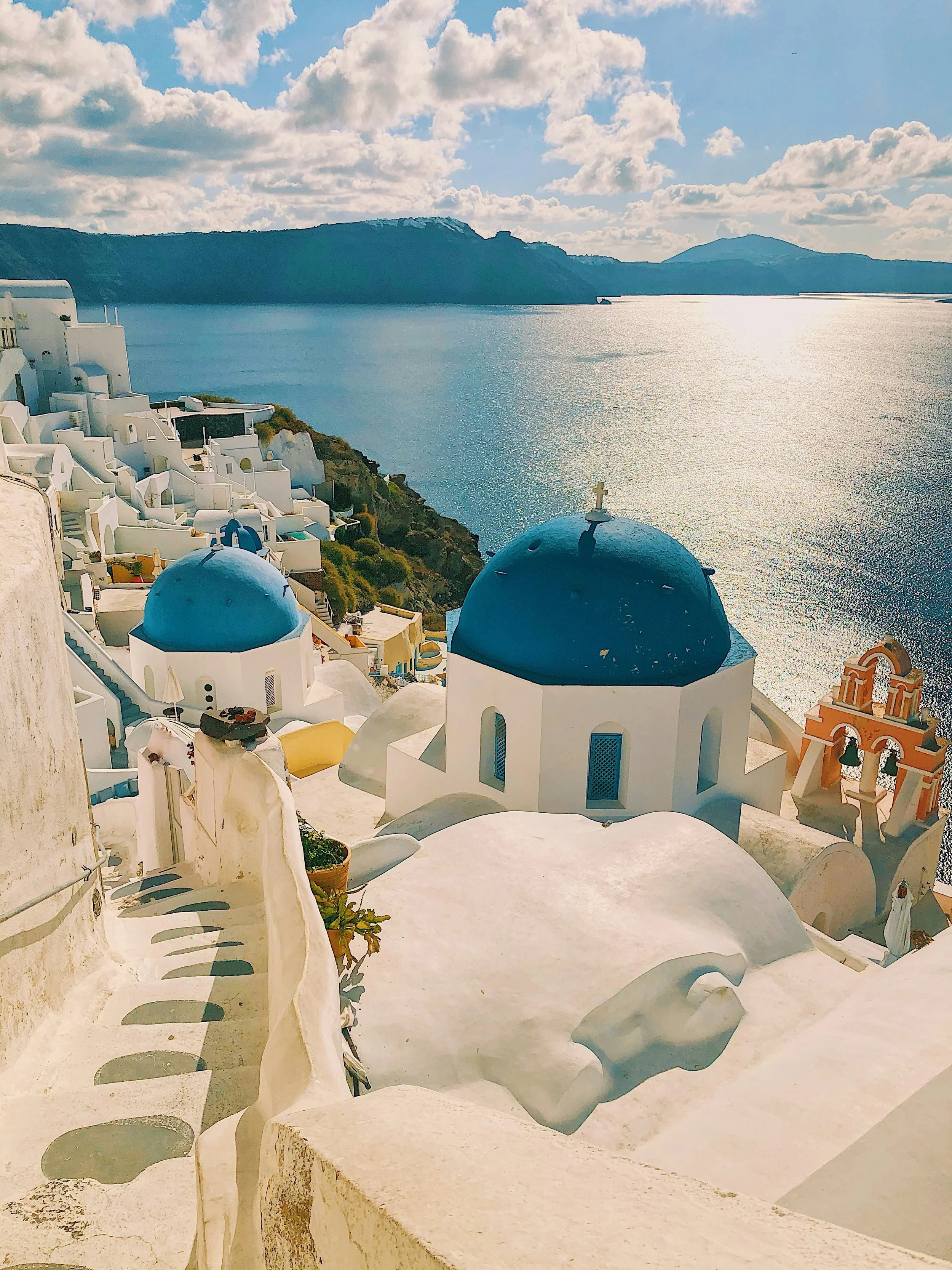 View of white buildings with blue domes on a hillside in overlooking a large body of water in Santorini, Greece, with a partly cloudy sky.