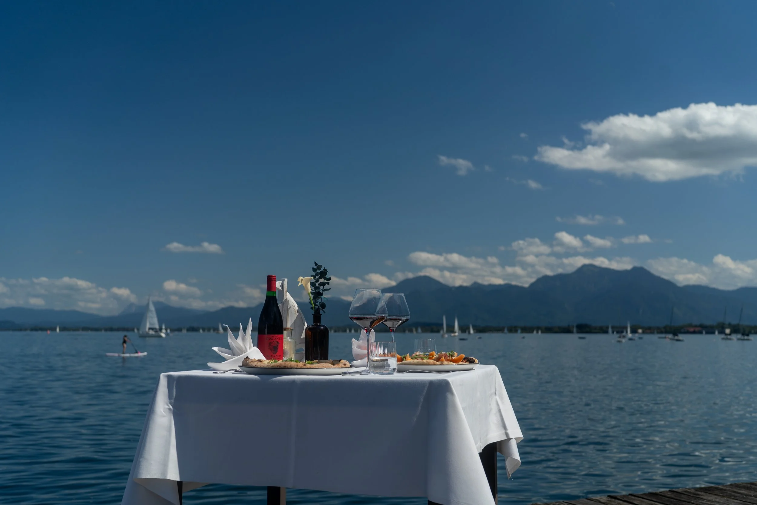 A table set with a bottle of wine, two glasses, and appetizers near a lake with sailboats and mountains in the background. Copyright Photography - Christoph Rutenkolk