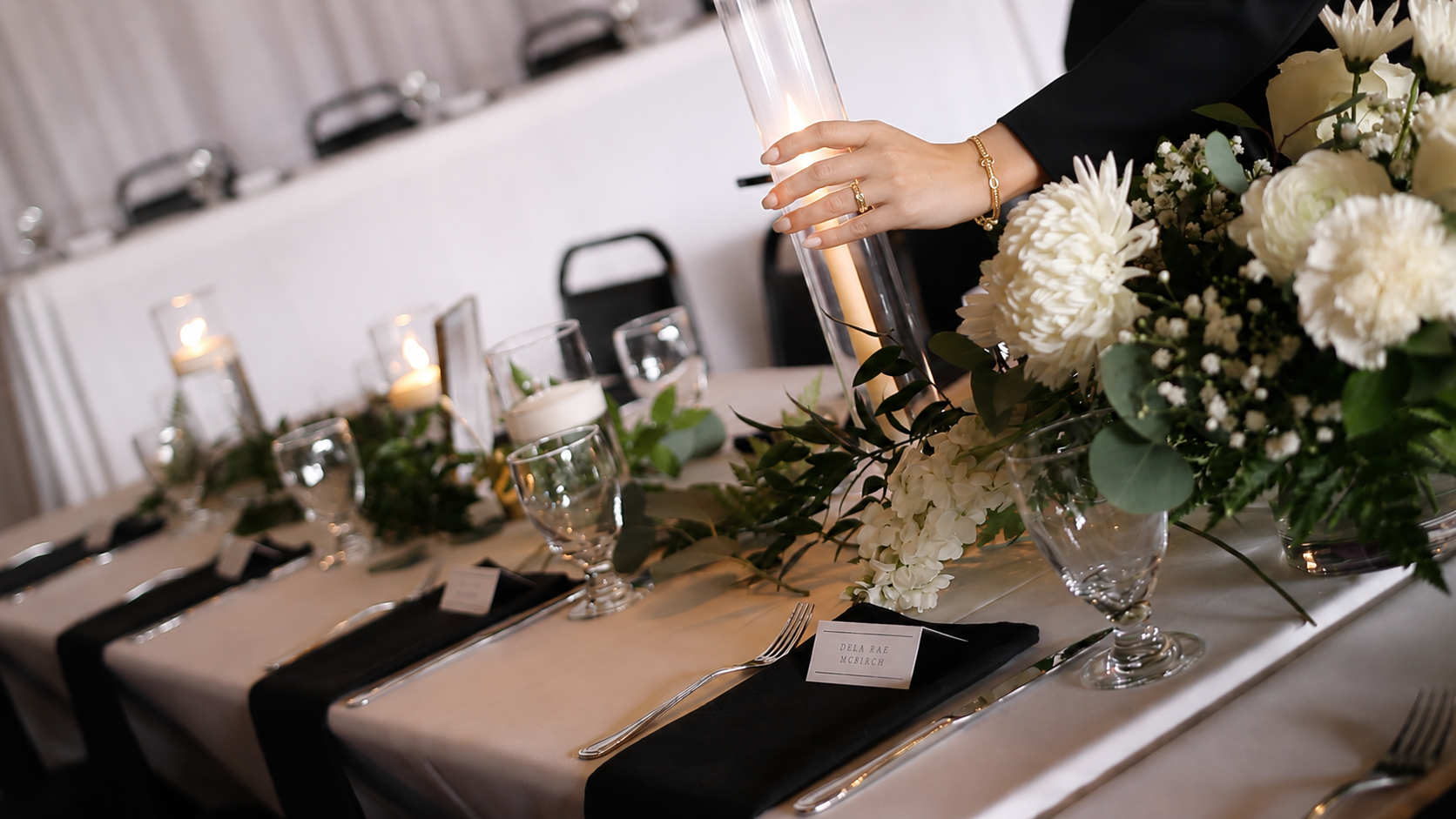 Event staff lighting candles on table scape featuring white florals, greenery, and black napkins