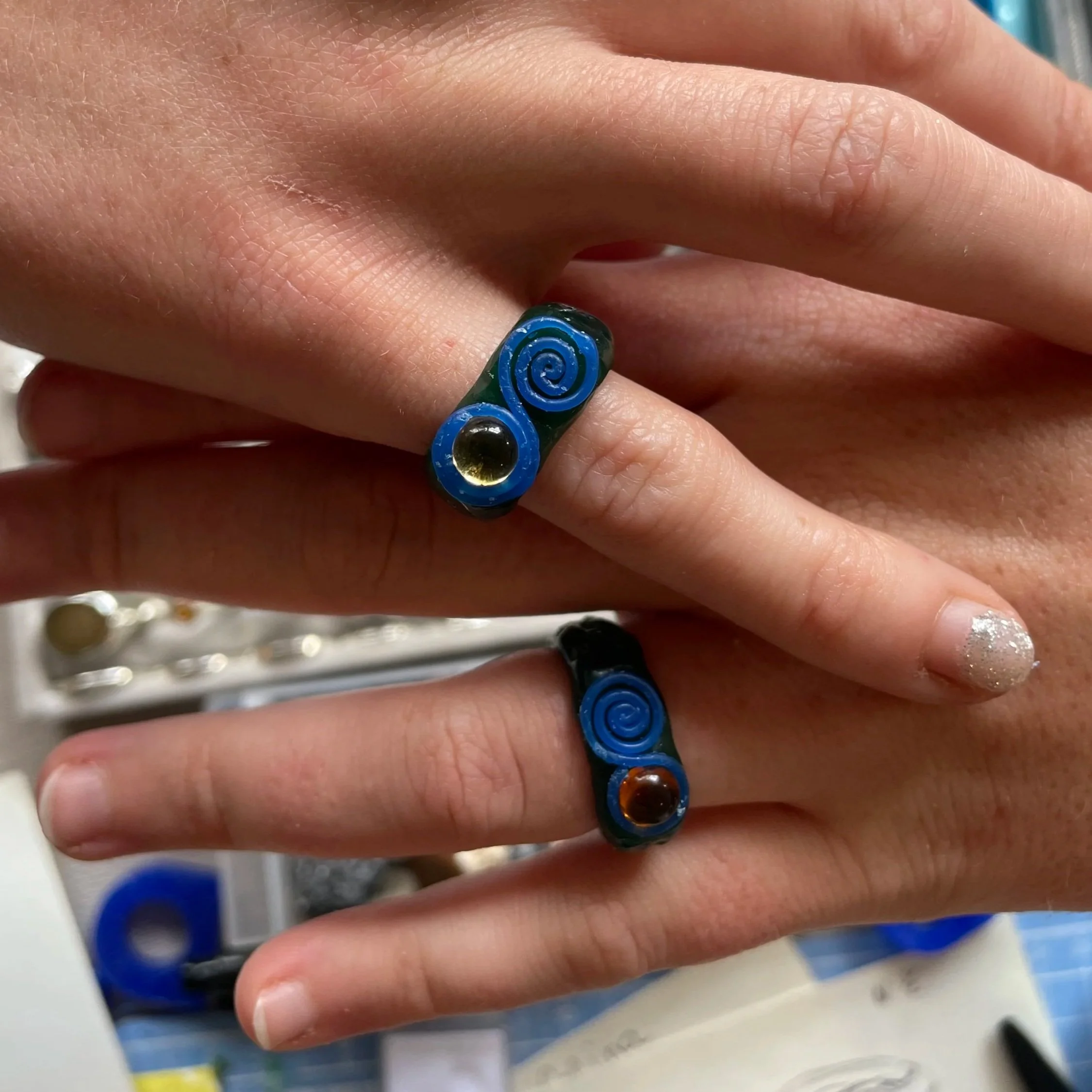 A hand with four rings on different fingers. The rings are silver with various design features including gemstones and textured patterns.