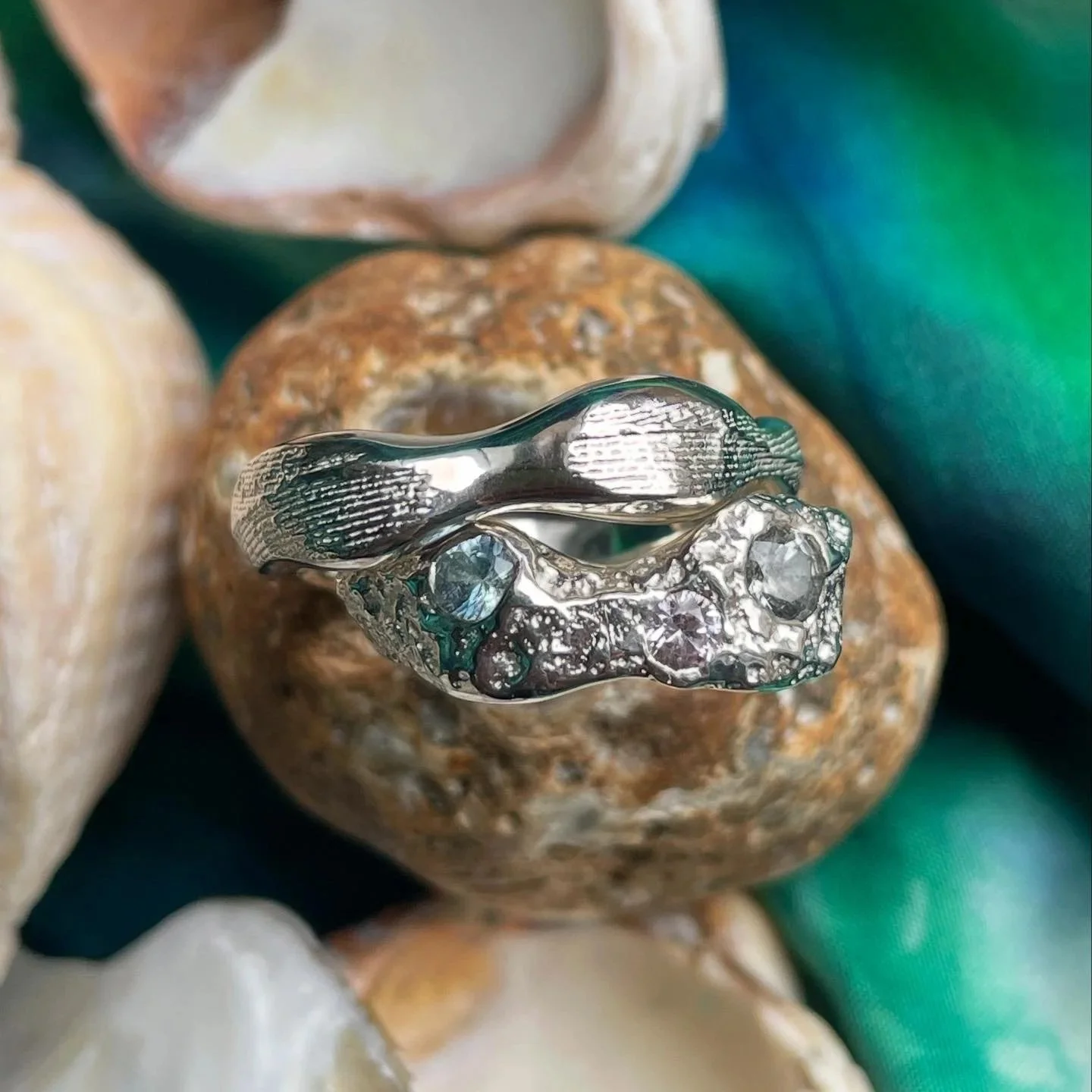 Close-up of a silver ring with embedded gemstones, resting on a brown stone with seashells and green fabric in the background.