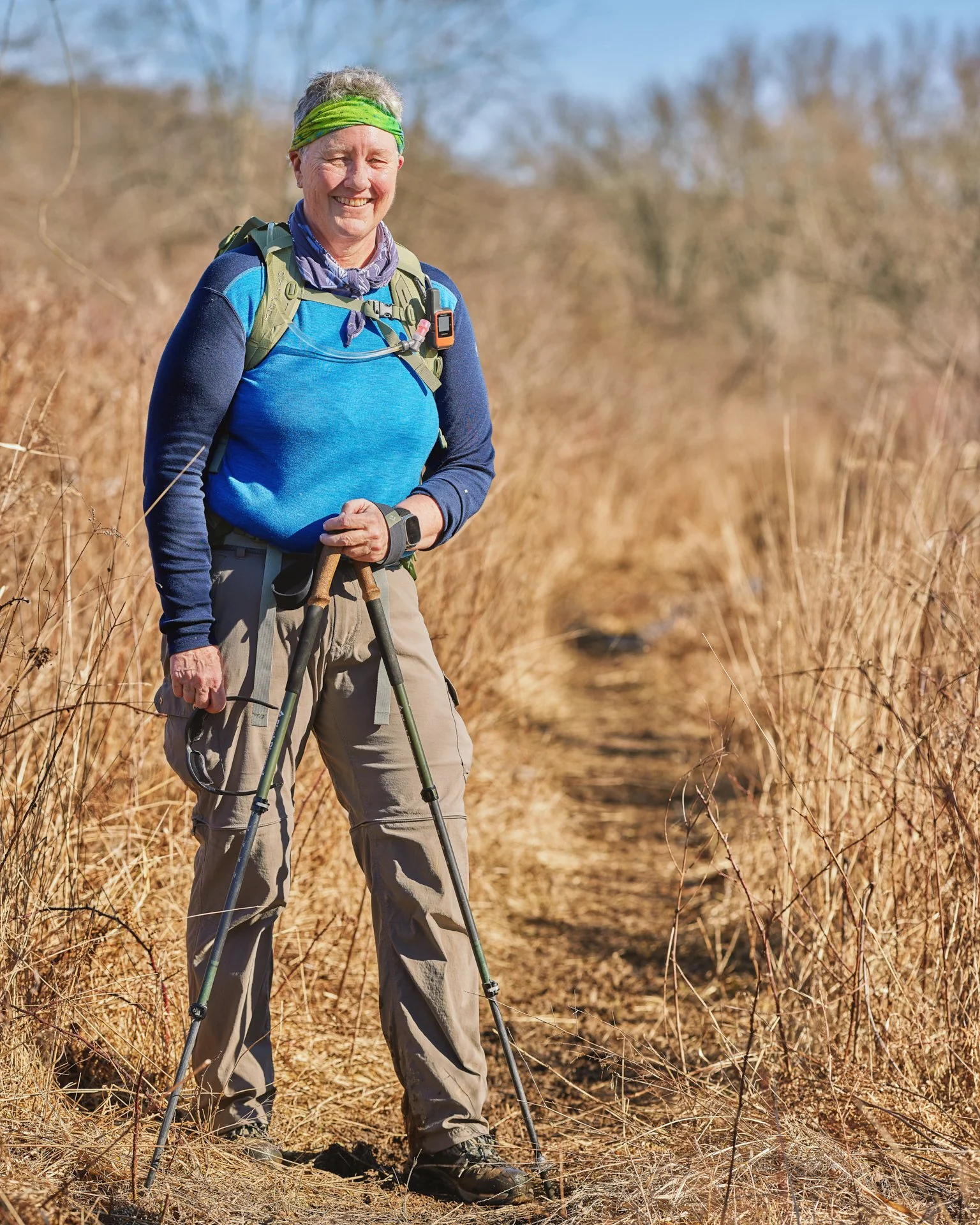 An elderly woman hiking on a dirt trail surrounded by dry grass on a sunny day.