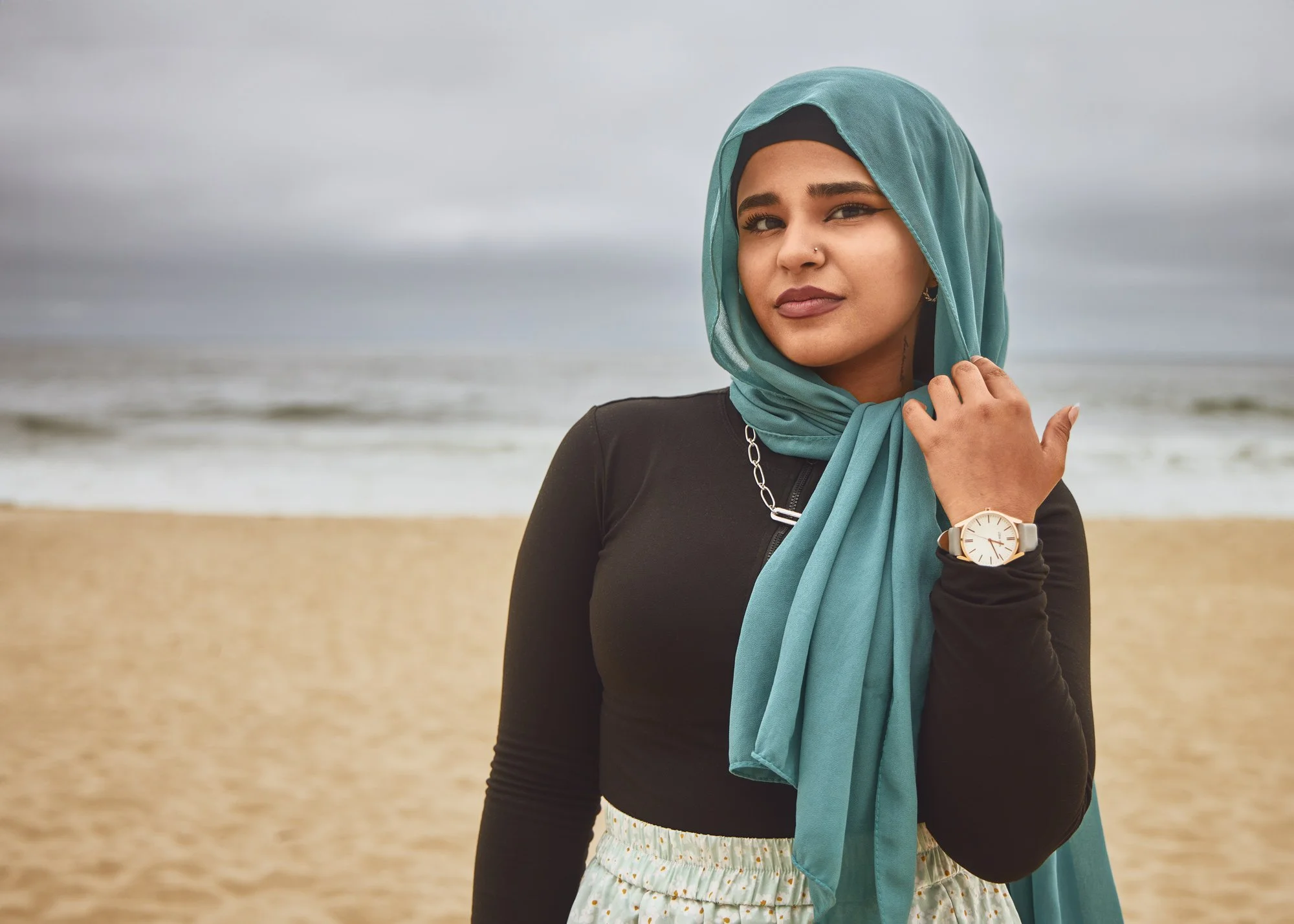 A woman wearing a teal headscarf, black top, and a watch, standing on a beach with sand and ocean in the background, overcast sky.