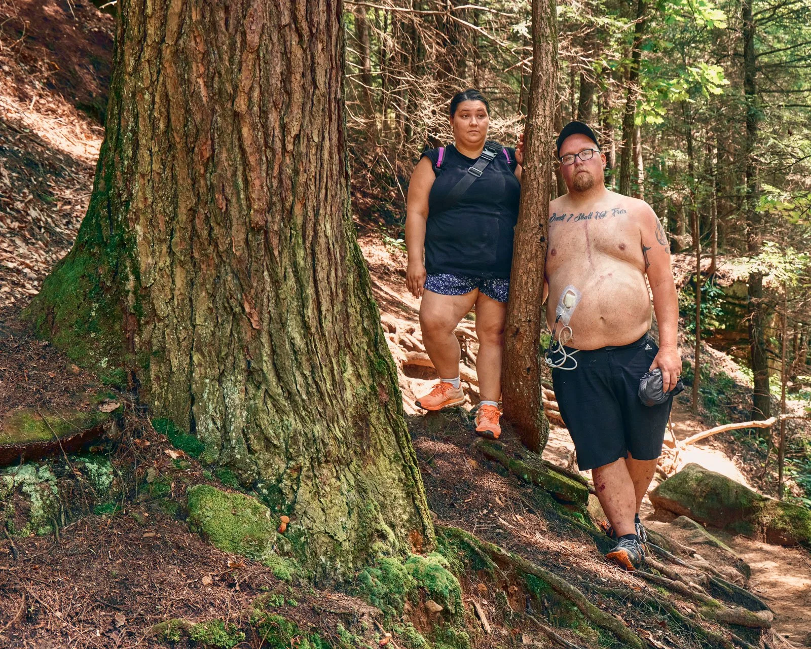 A woman and a shirtless man stand on a forest trail surrounded by trees, with the man holding a camera and the woman leaning against a tree.