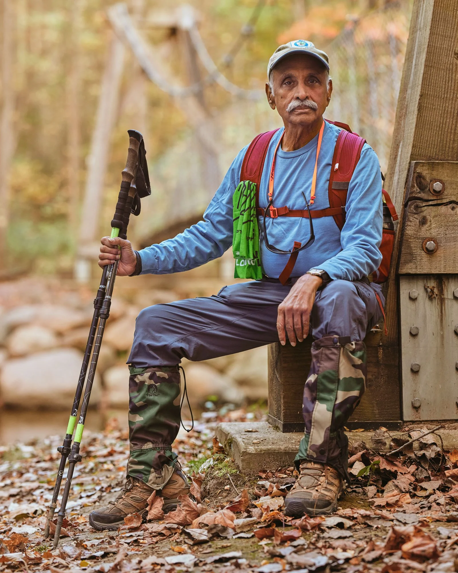 An older man in hiking gear sitting on a wooden structure in a forest with fallen leaves, holding trekking poles, wearing a blue long-sleeve shirt, camouflage gaiters, and a red backpack.