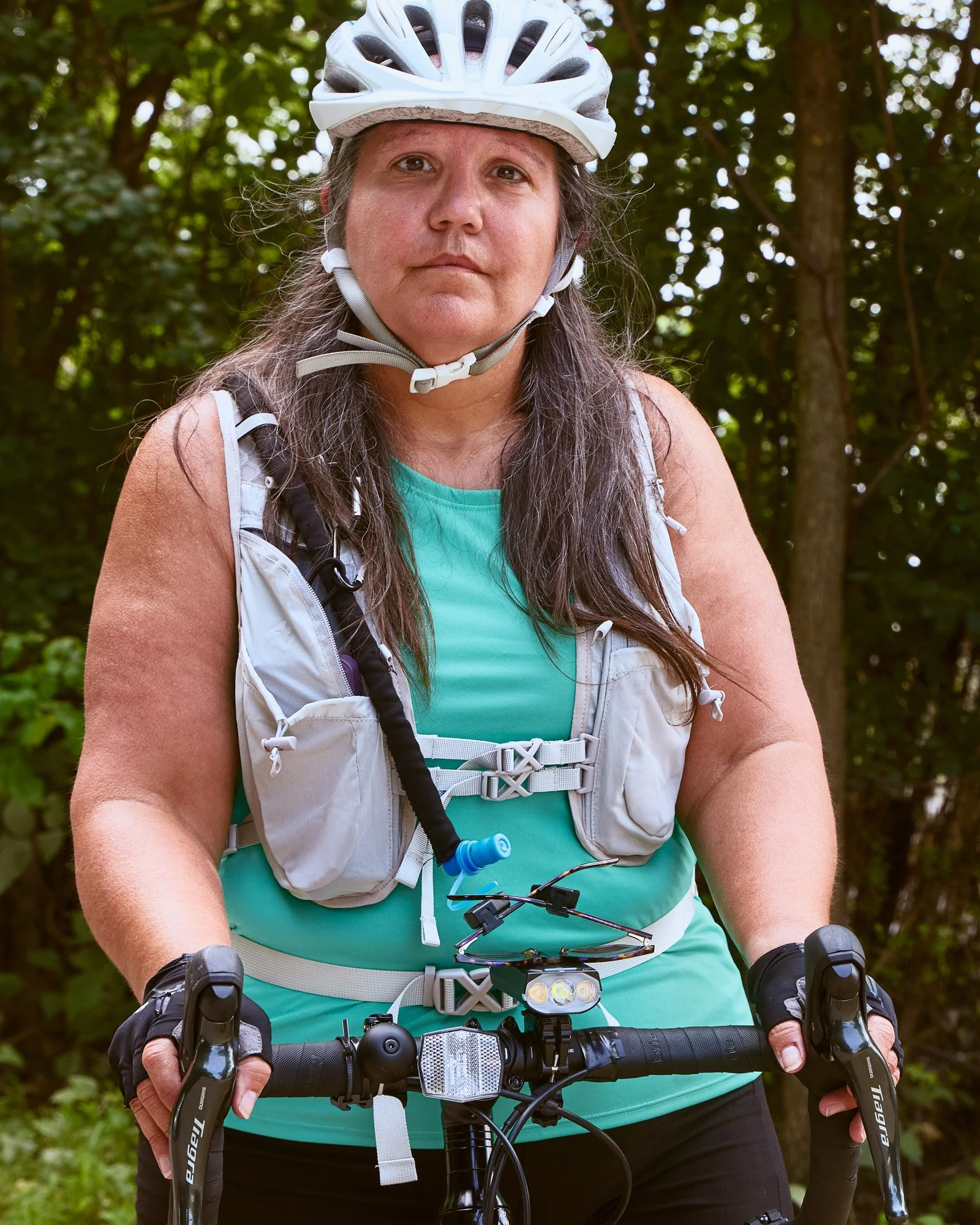 A woman wearing a white bicycle helmet and riding gear, holding a bike handlebar, standing outdoors with trees in the background.