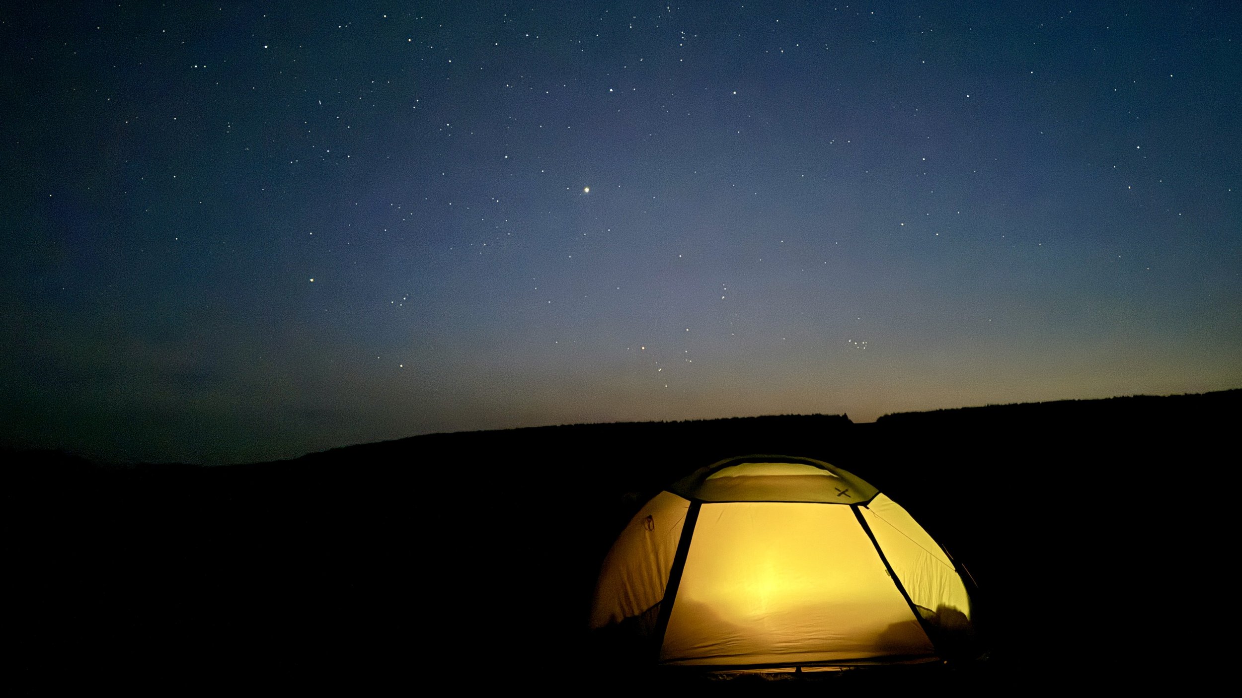A glowing tent under a starry night sky in a dark landscape.