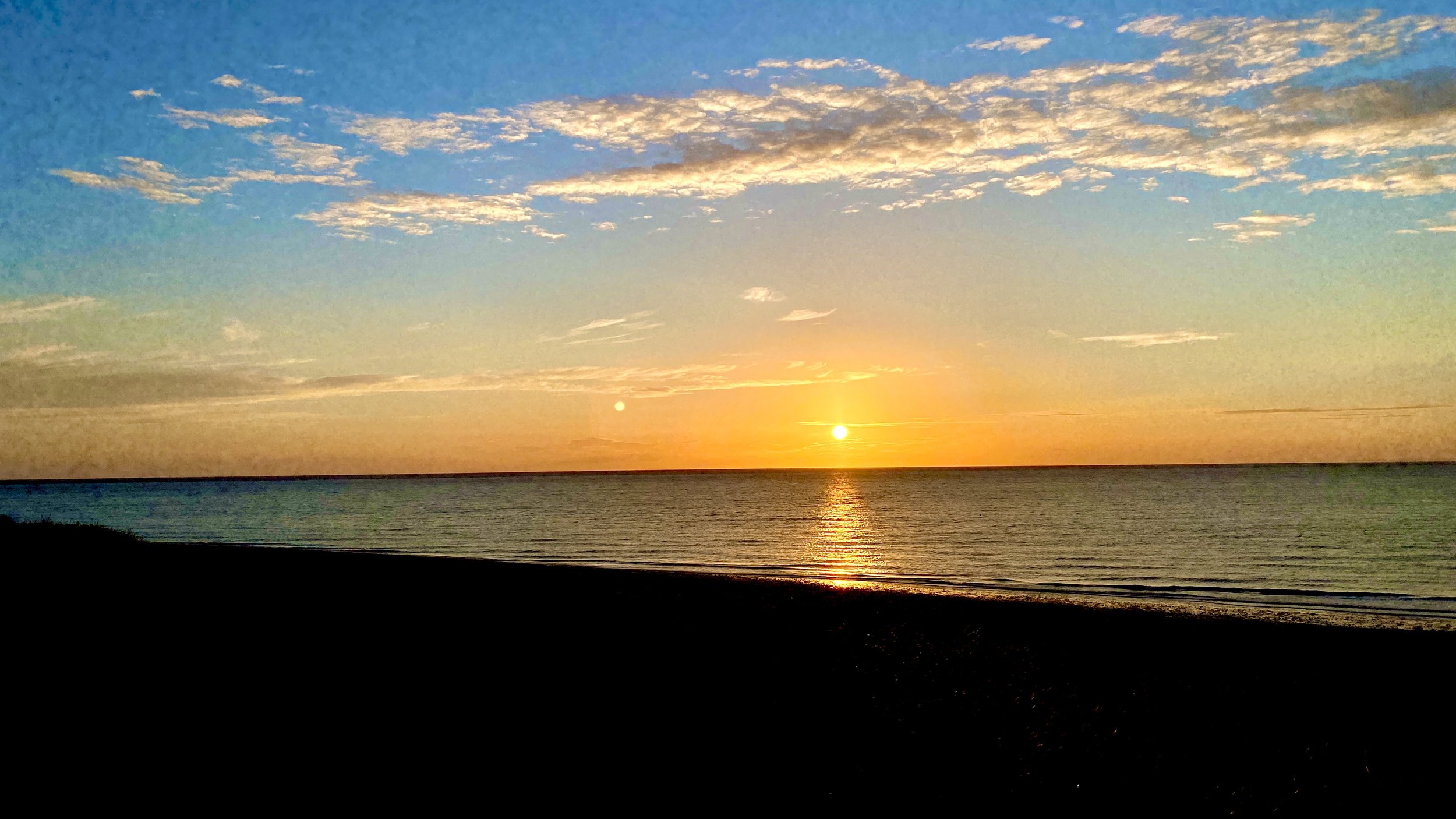 Sunset over the ocean with a sandy beach in the foreground, colorful sky with clouds.