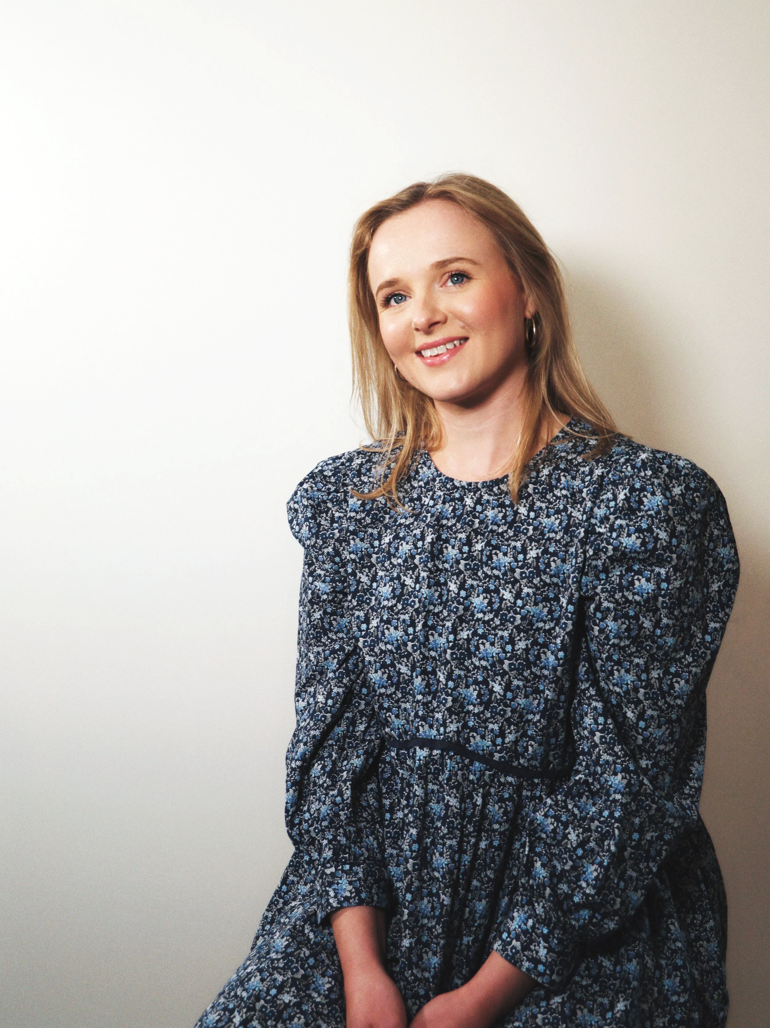 A woman with blonde hair wearing a blue floral dress, smiling while sitting against a plain wall.