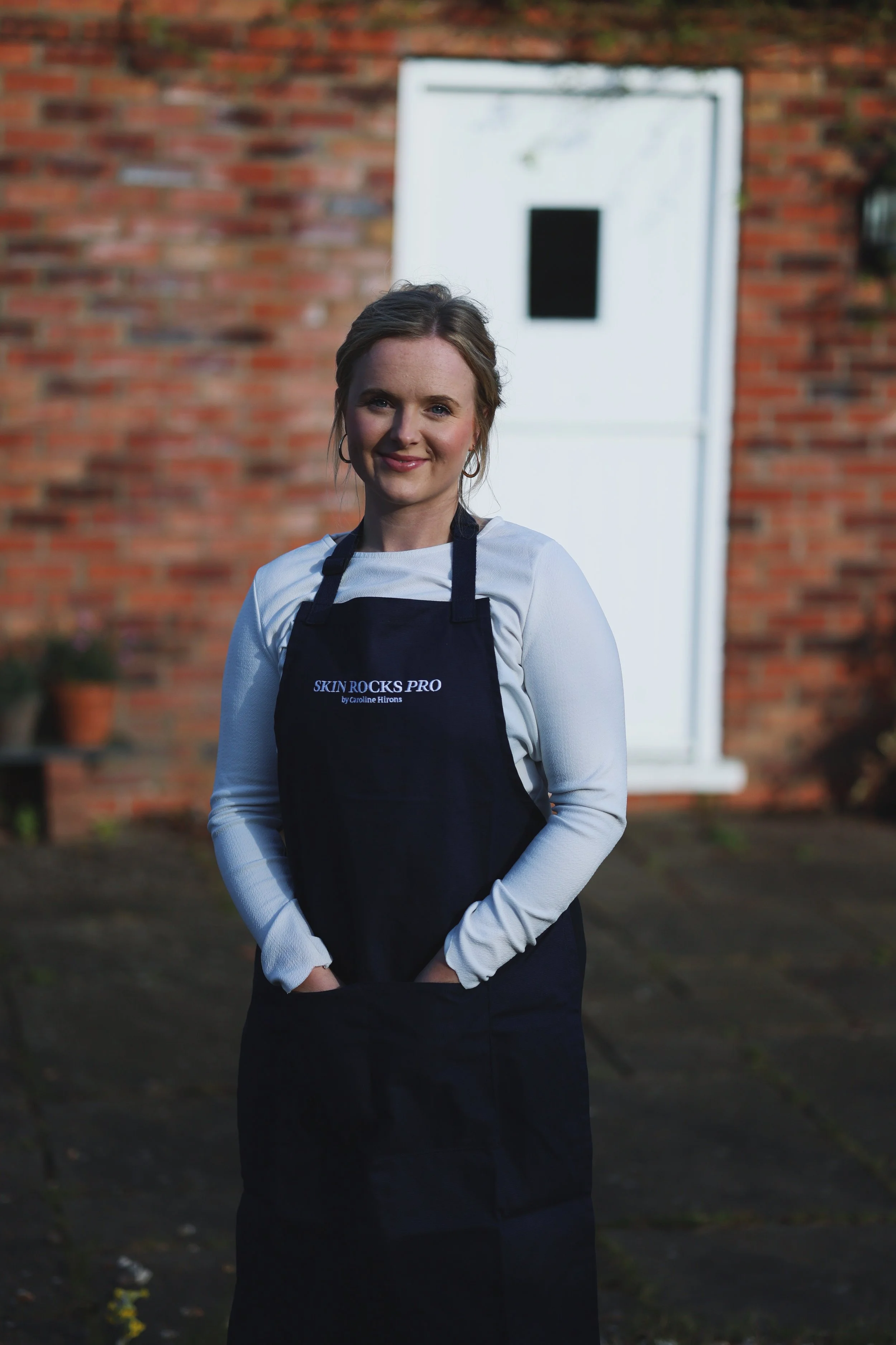 A woman standing outdoors on a brick patio, wearing a navy apron with the text "SKIN ROCKS PRO" and a white long sleeve shirt, smiling at the camera.