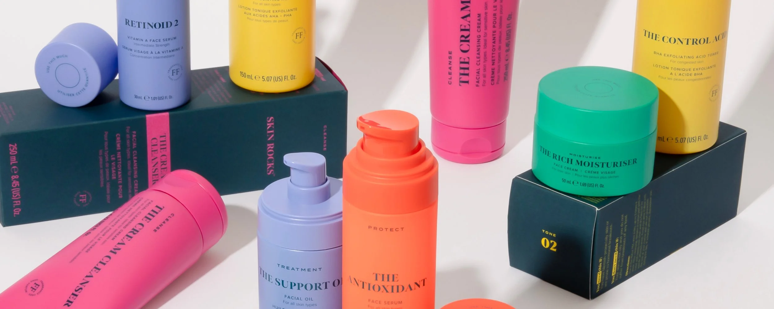 Collection of colorful skincare products, including bottles and jars, arranged on boxes and a white surface. The products are in shades of purple, pink, yellow, orange, and green.