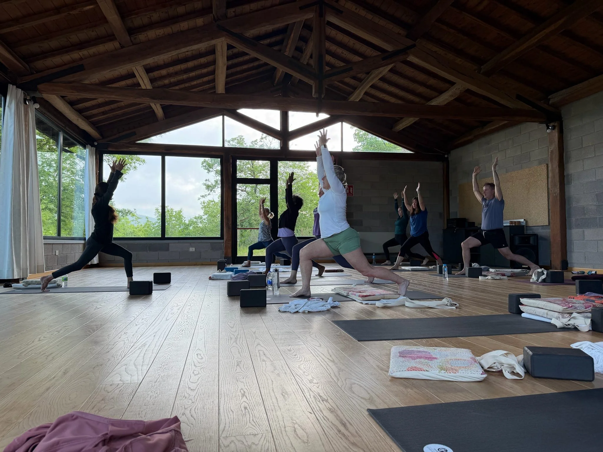 Group of people practicing yoga in a spacious studio with large windows and wooden ceiling beams. They are in a pose with arms raised and legs bent, standing on yoga mats with blankets and water bottles nearby.