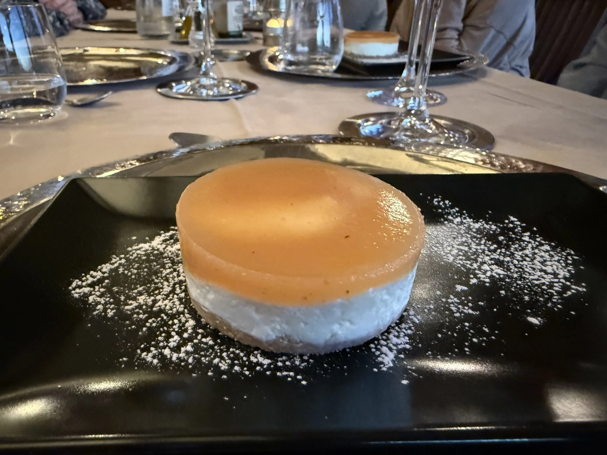 A dessert on a black plate topped with powdered sugar and a small dollop of whipped cream, with a dinner table in the background.