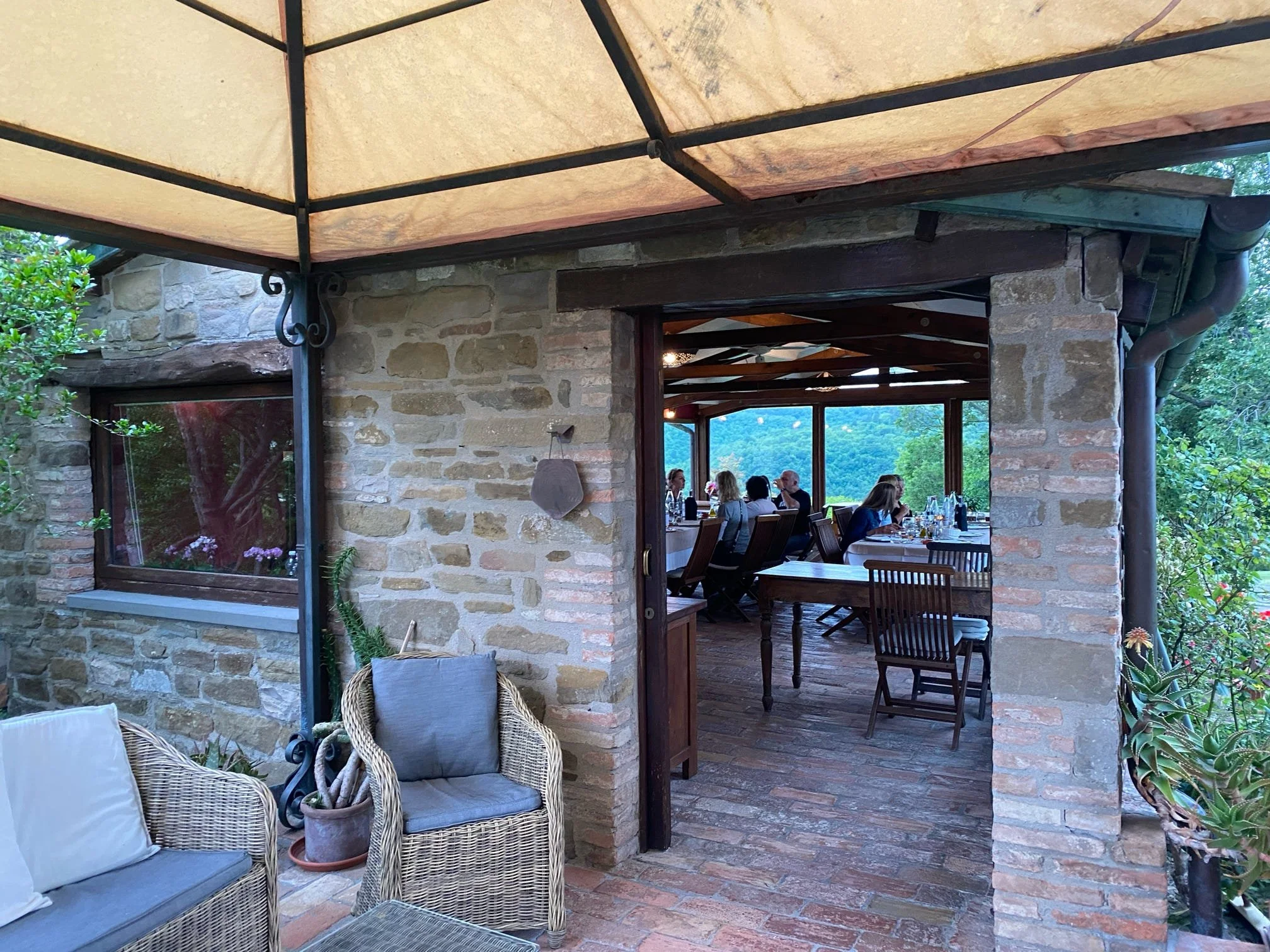 Outdoor restaurant or patio area with a stone wall, wooden beams, a large window, and seating including wicker chairs with cushions. There are people dining inside, with a scenic view of trees and mountains in the background.