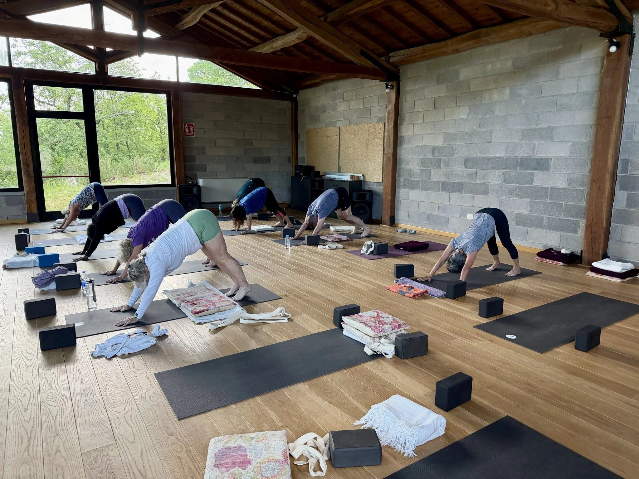 A group of people participating in a yoga class in a spacious room with large windows, wooden ceiling, and concrete walls. They are in a downward dog pose on yoga mats, with various belongings placed beside them.