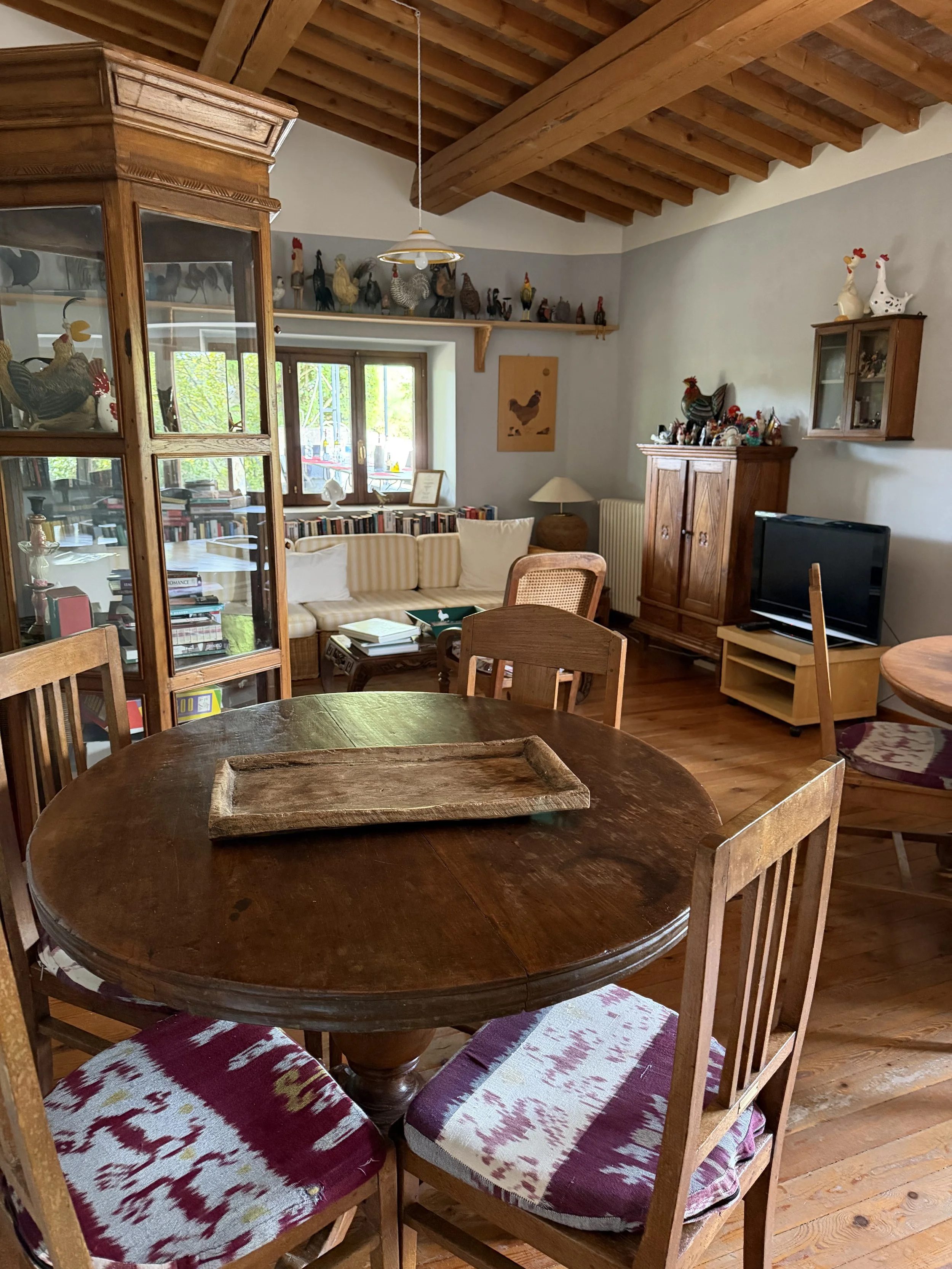 A cozy living room with wooden ceiling beams, a round dining table with four chairs, and a cabinet filled with decorative items and books. There is a window with a view of trees outside, and a couch with striped pillows.