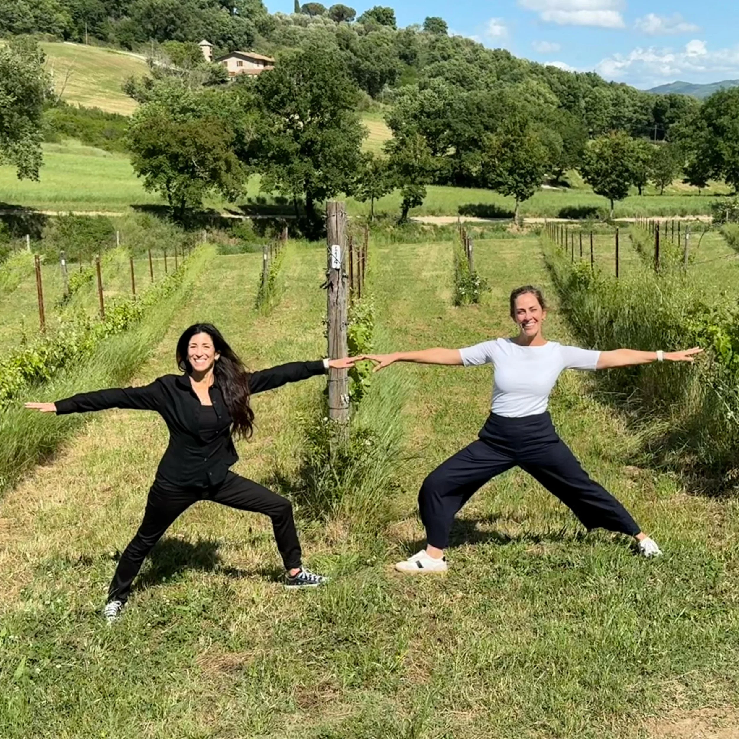 Two women are standing in a vineyard, smiling and touching each other with their arms outstretched. The vineyard has rows of grapevines and is surrounded by green trees and hills under a blue sky with some clouds.