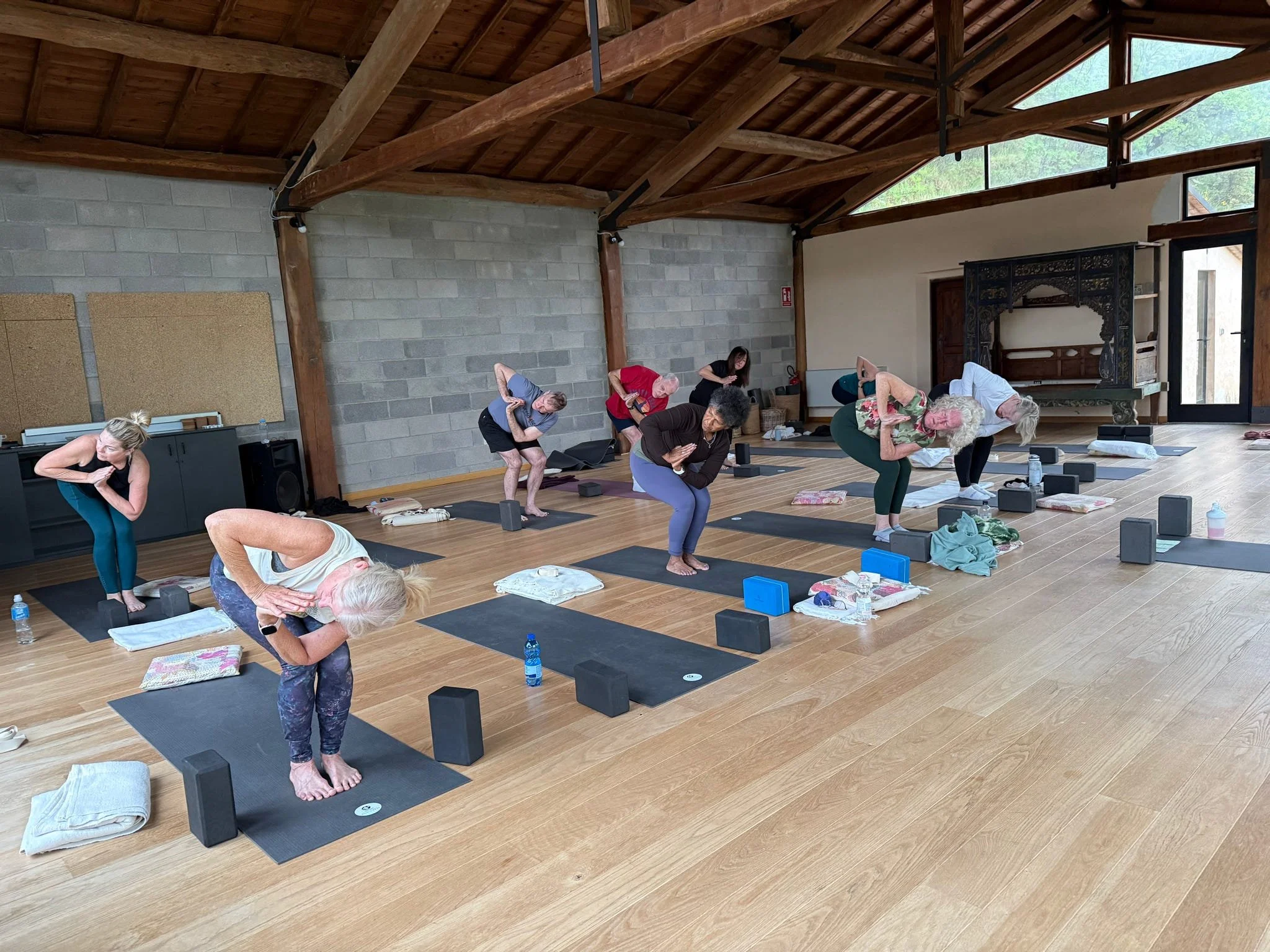 A group of people participating in a yoga class in a spacious studio with wooden beams and large windows, practicing poses on yoga mats with props.