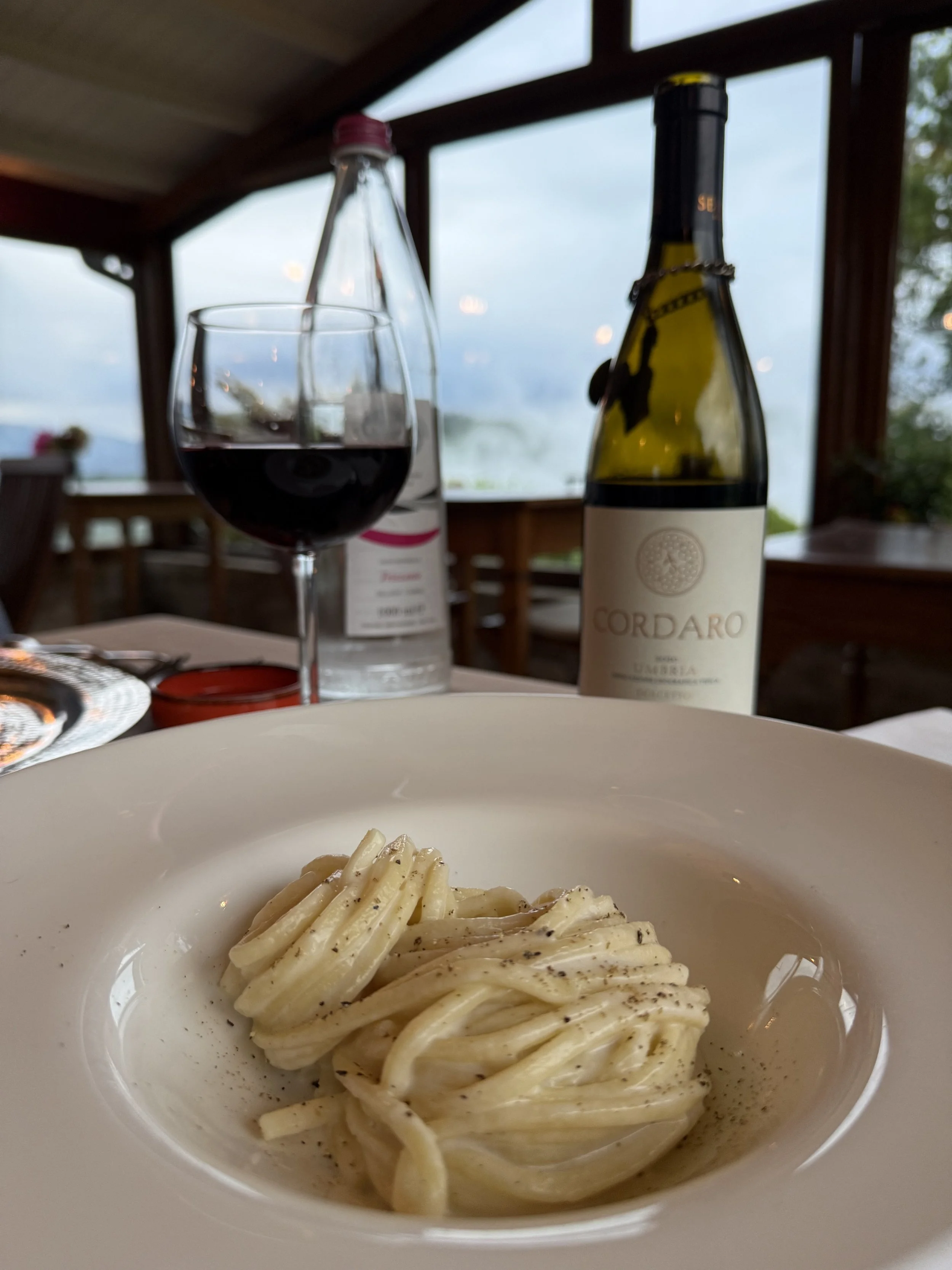 A white bowl filled with creamy pasta, possibly fettuccine or linguine, topped with black pepper. In the background, there is a glass of red wine, a bottle of white wine labeled 'Cordaro', and a perch of water on a dining table in a room with large w