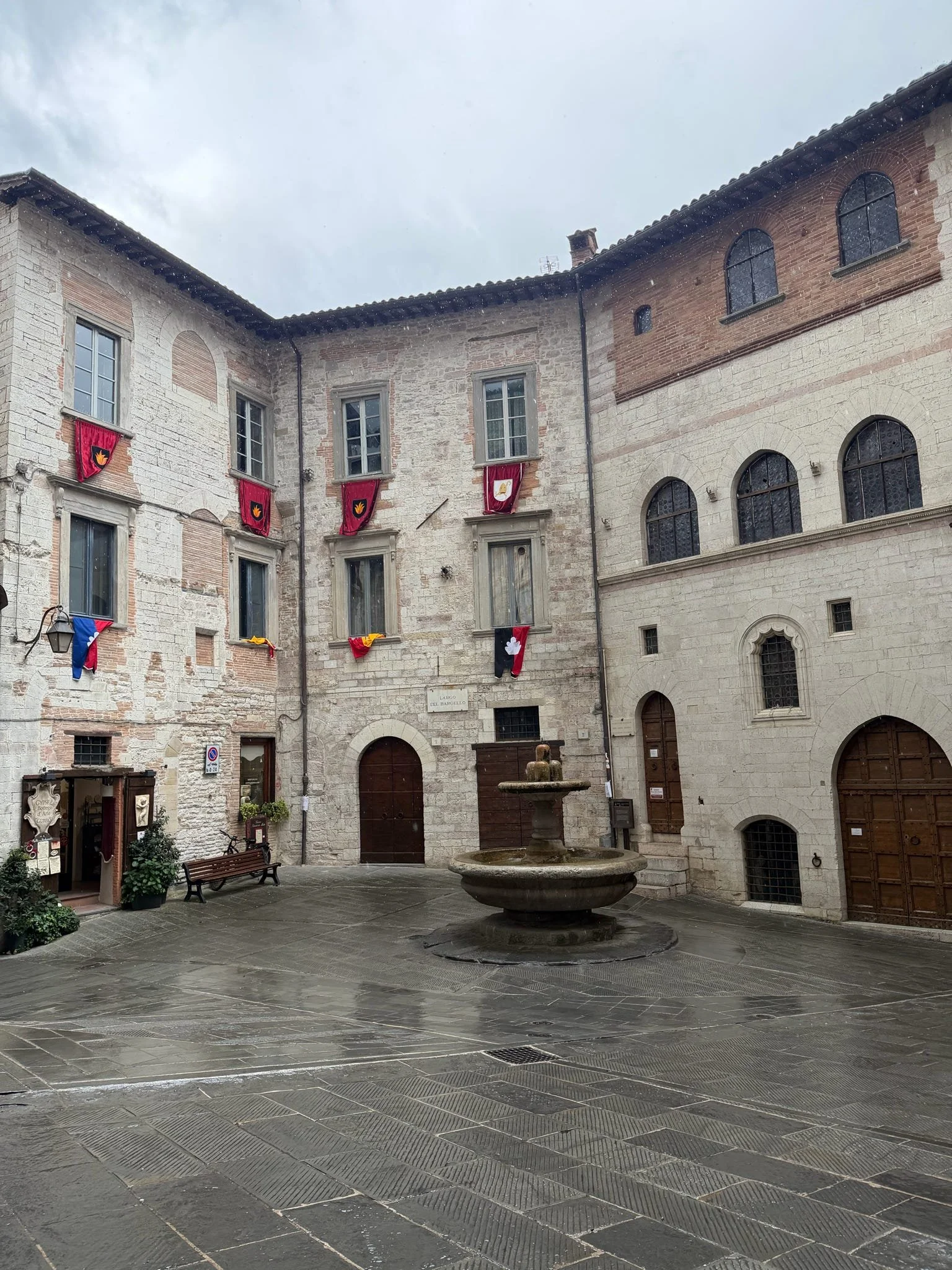 A small European-style courtyard with a stone fountain in the center, surrounded by historic stone and brick buildings with arched windows, a bench, and hanging banners on the walls.