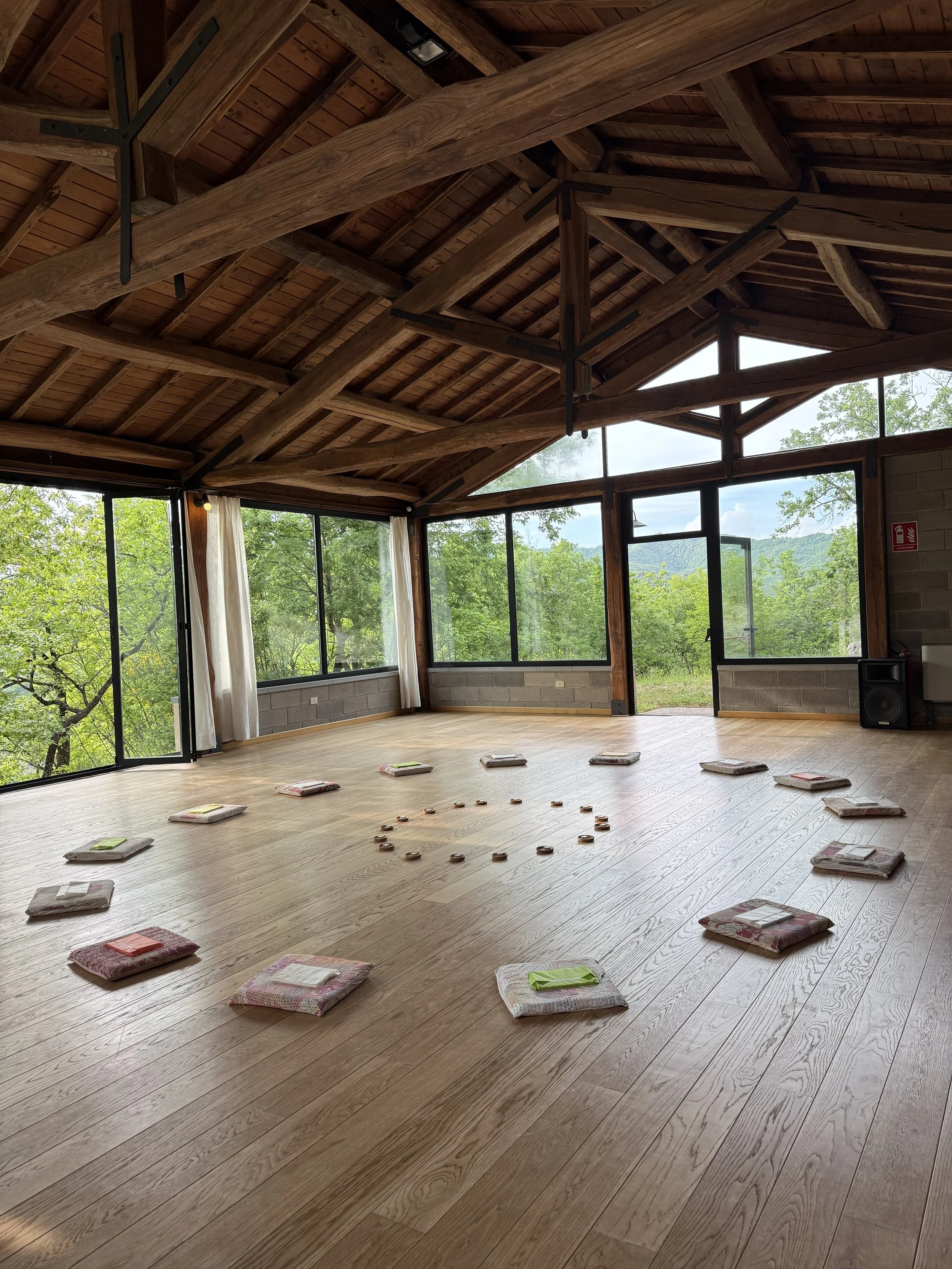An indoor space with wooden floors, large windows, and a wooden ceiling with exposed beams. Cushions are arranged in a circle on the floor for a group activity, and there is outdoor greenery visible through the windows.