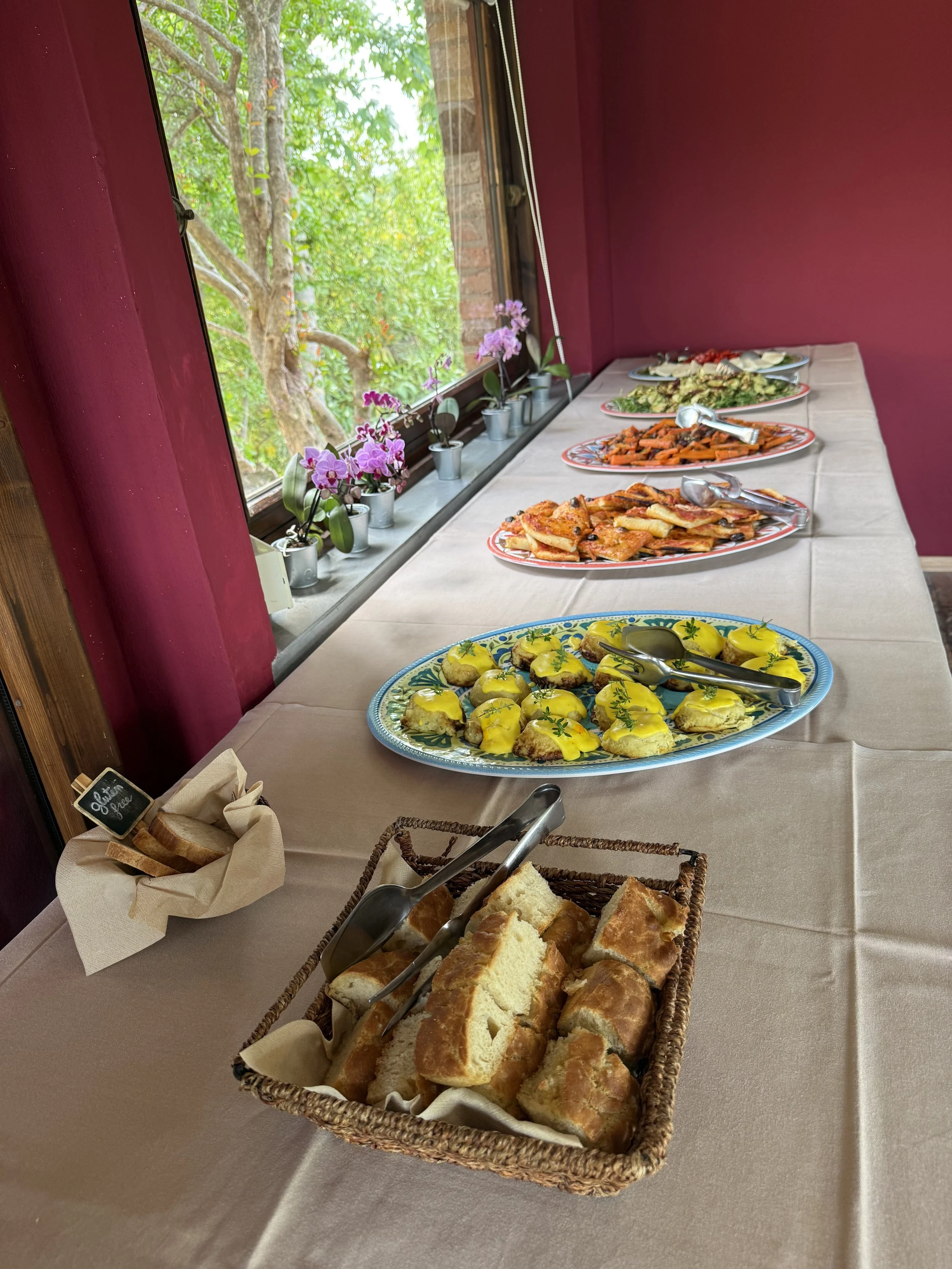 A buffet table with various dishes including bread, pastries, and appetizers, set on a beige tablecloth near a window with pink walls and potted orchids outside.