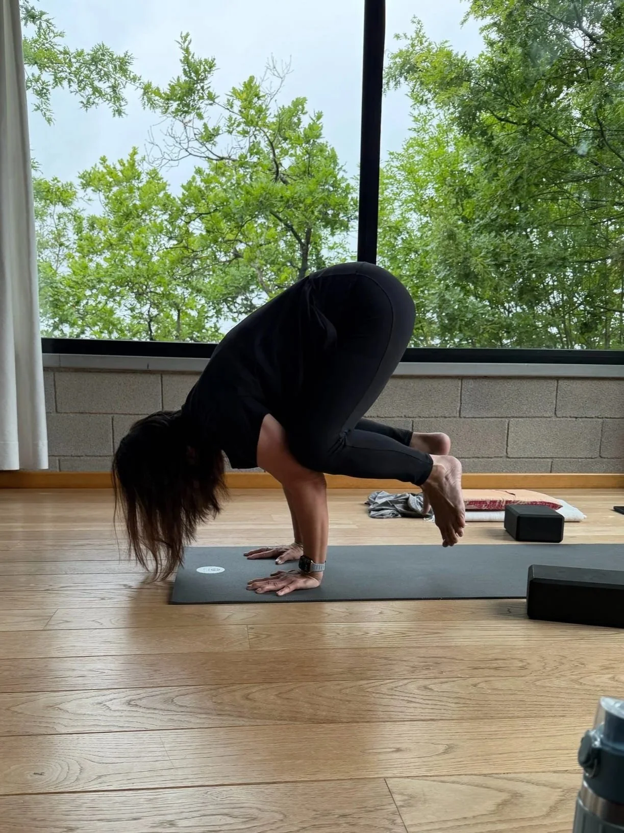 A woman practicing yoga indoors on a black mat, performing a crow pose with her hands on the mat and knees resting on her upper arms, with a large window showing green trees outside.