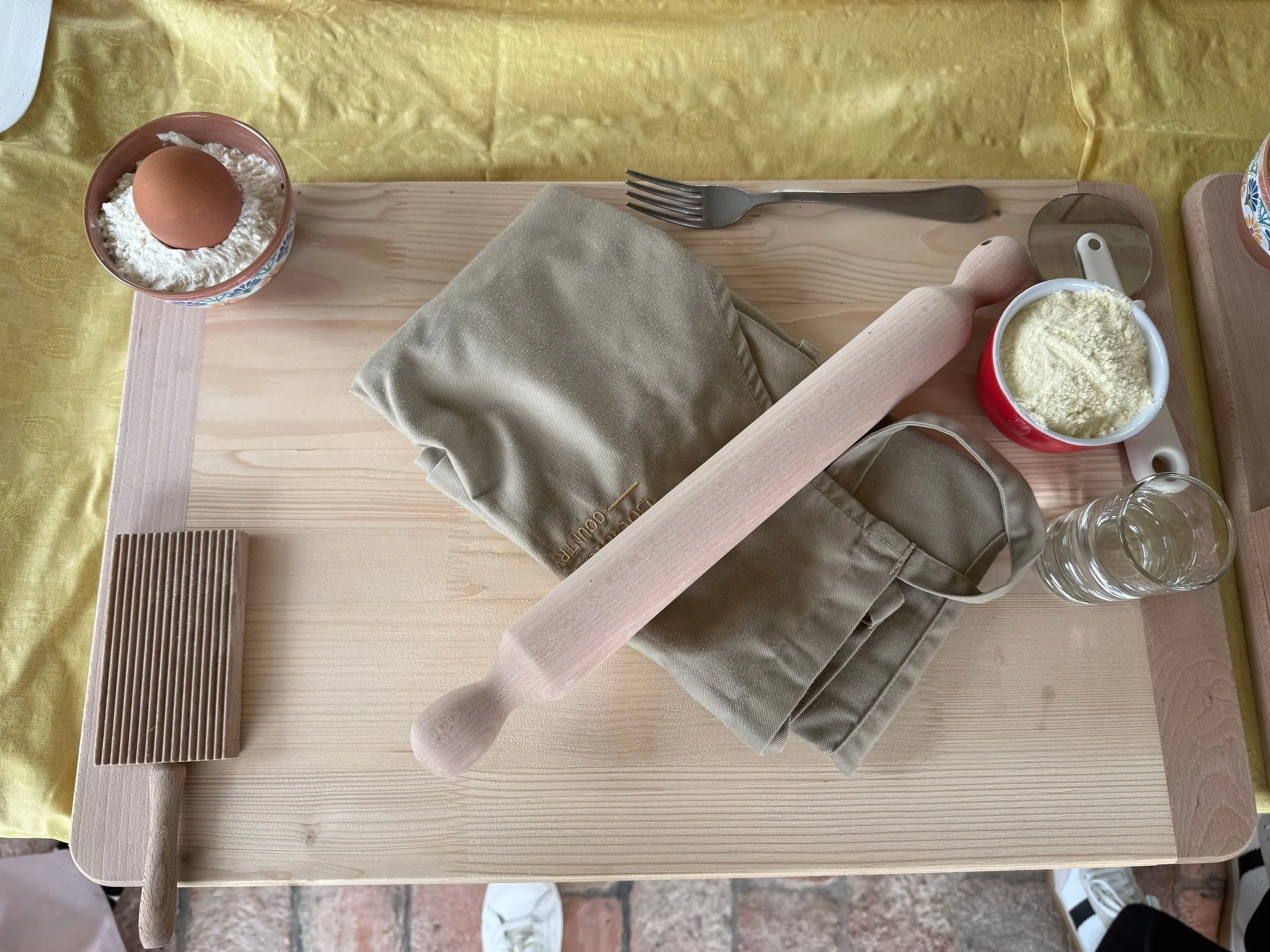 A wooden table with cooking ingredients and utensils, including a bowl of flour with an egg on top, a cup of flour, a rolling pin, a fork, a small mirror, a glass, and a beige cloth napkin, with a yellow tablecloth underneath.