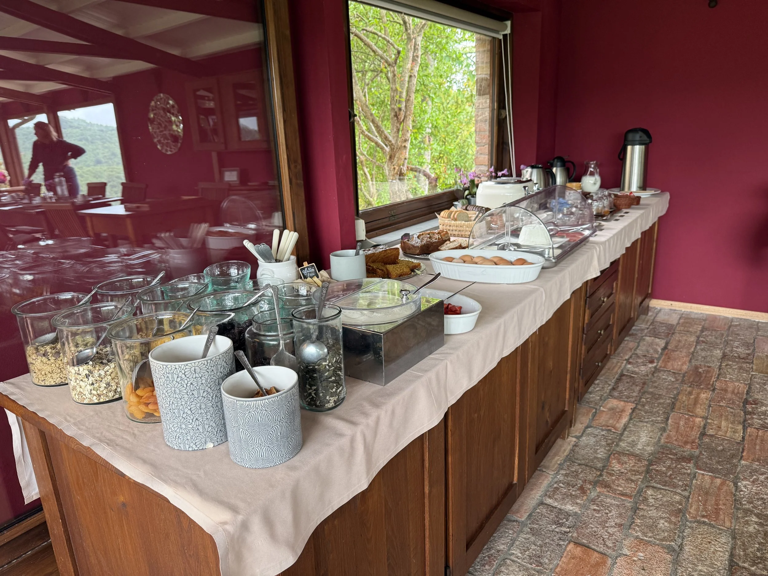 Buffet table set up with various cereals, eggs, baked goods, and beverages in a room with a window overlooking greenery.