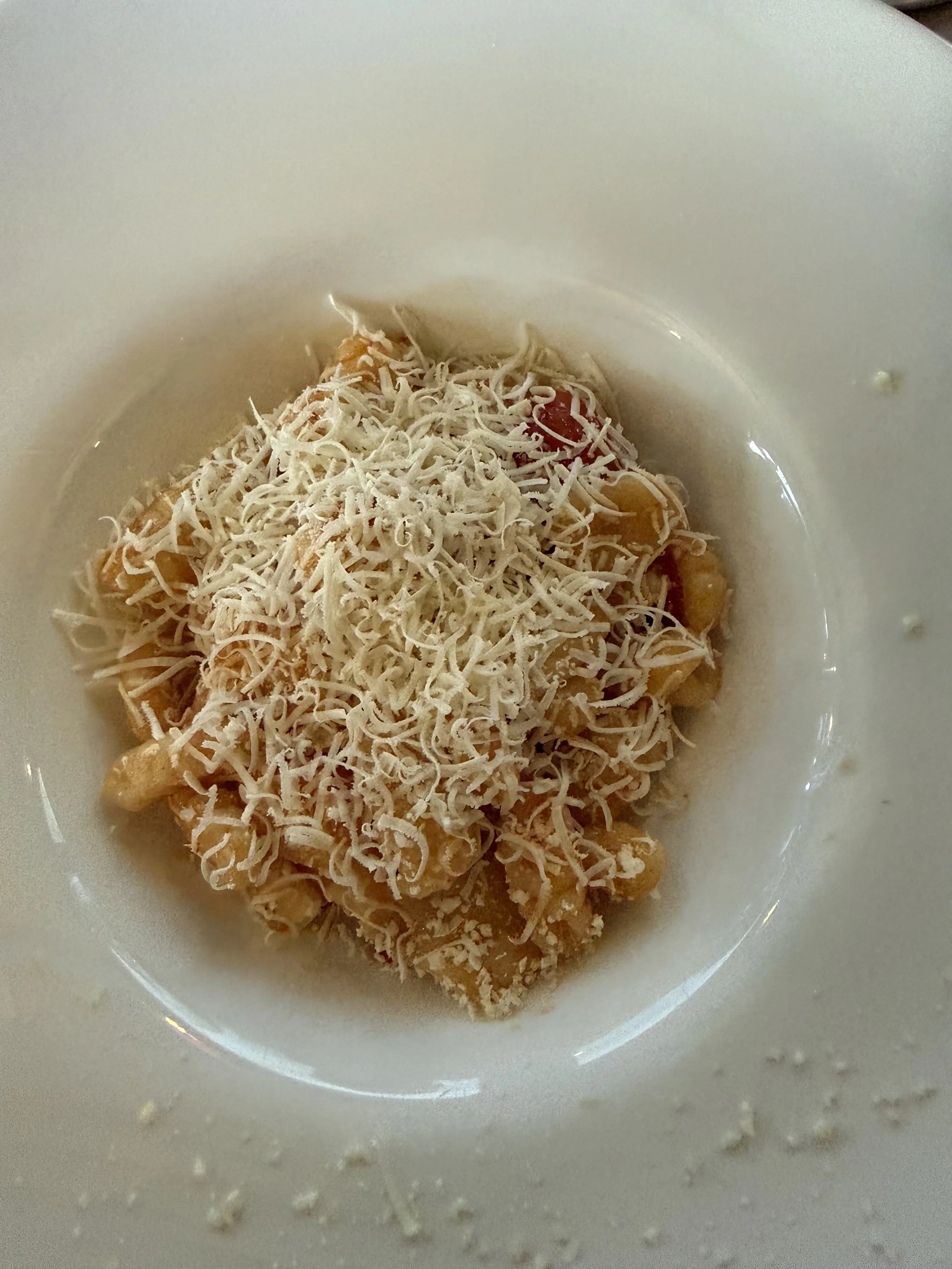 A white bowl filled with a dish topped with shredded cheese, which also contains cooked pasta, cherry tomatoes, and possibly other ingredients.
