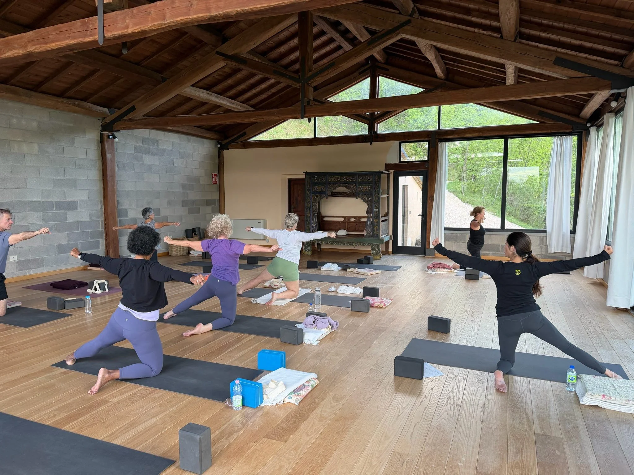 A group of people participating in a yoga class inside a room with large windows, wooden ceiling beams, and natural light, practicing yoga poses on mats with blocks, water bottles, and towels nearby.