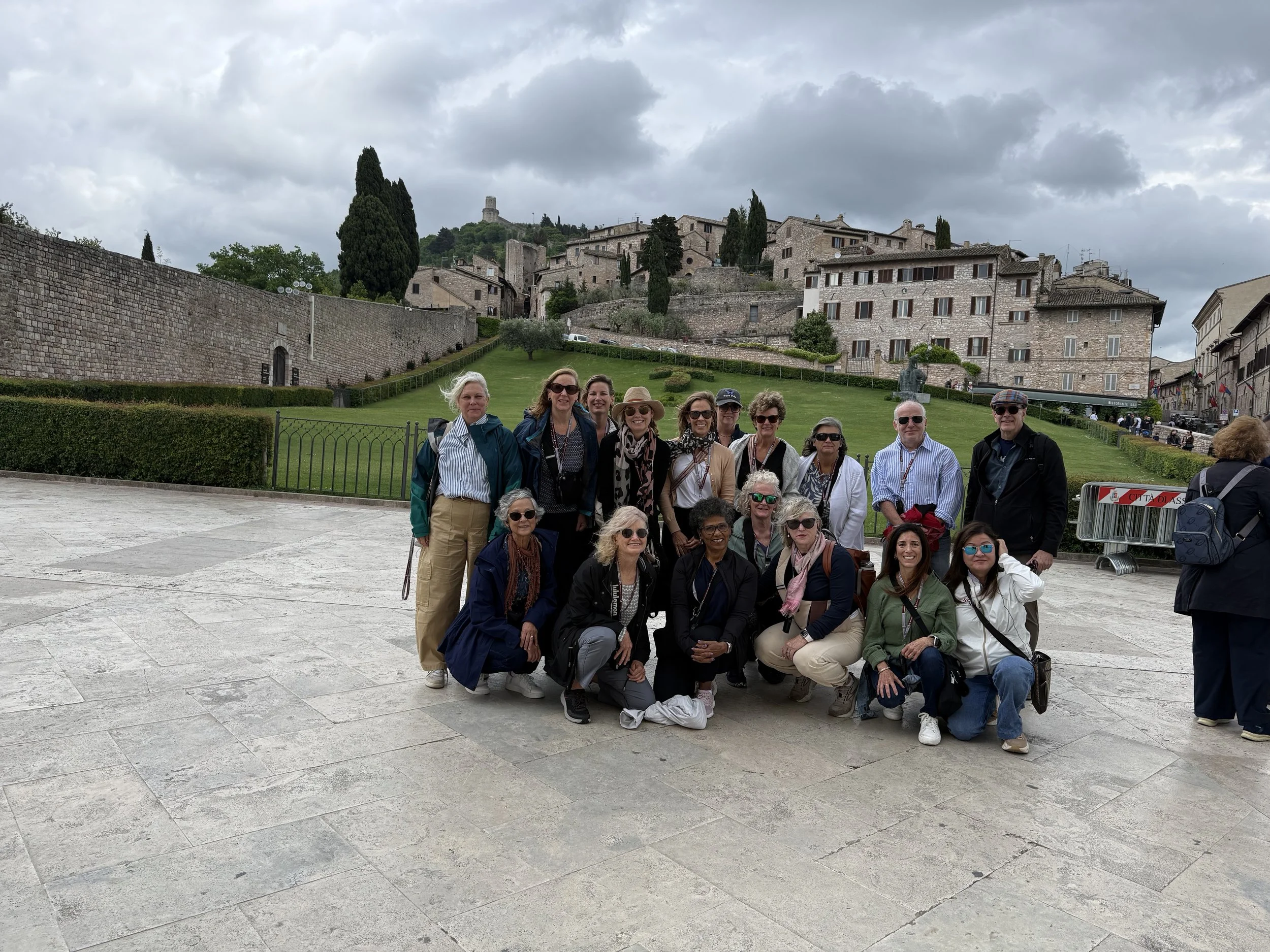 A group of tourists posing for a photo in front of a historic hilltop town with stone buildings, cypress trees, and a cloudy sky.
