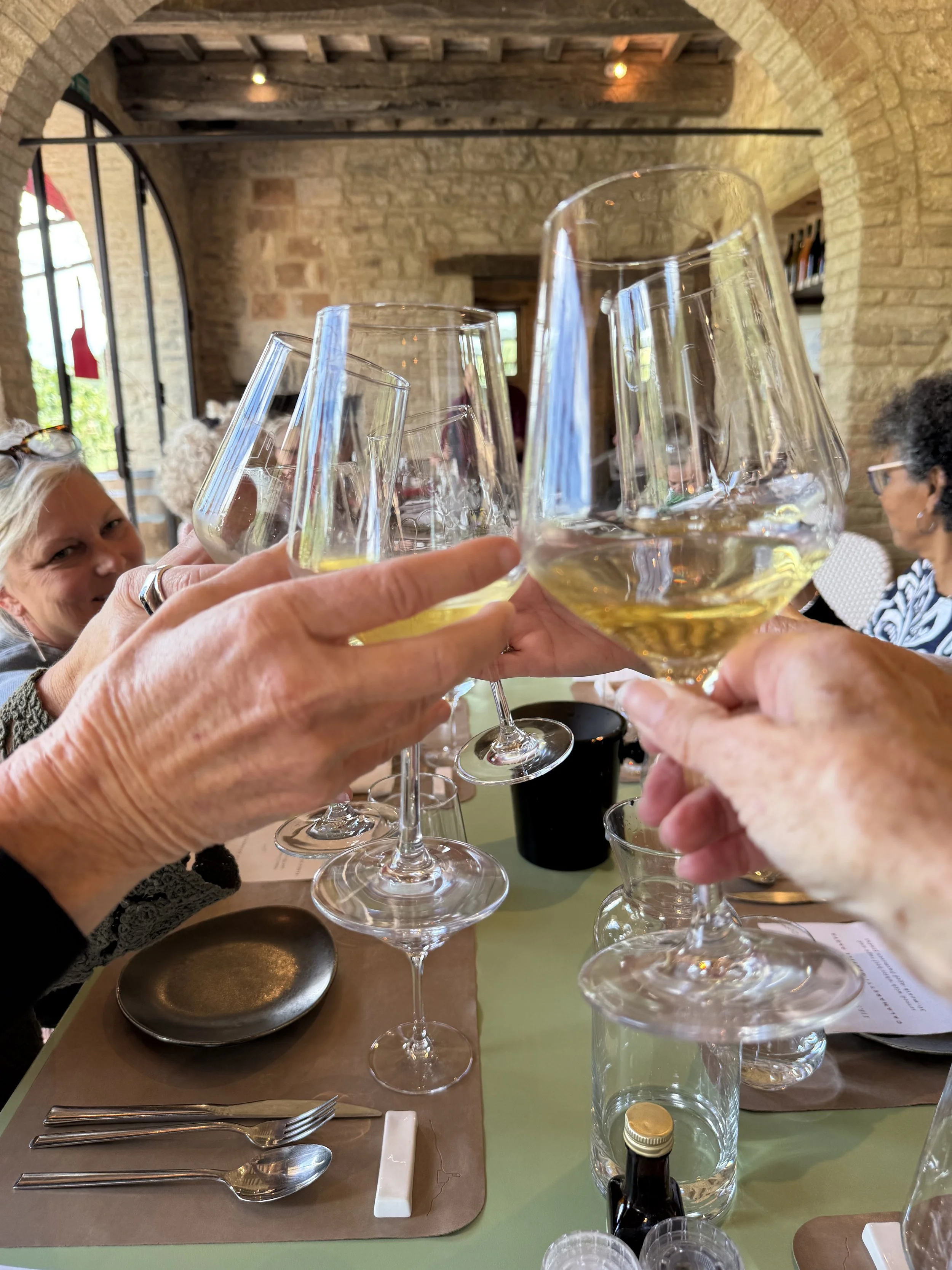Close-up of multiple glasses of white wine being toasted together in a restaurant with brick walls and wooden ceiling beams. Several people are visible in the background.