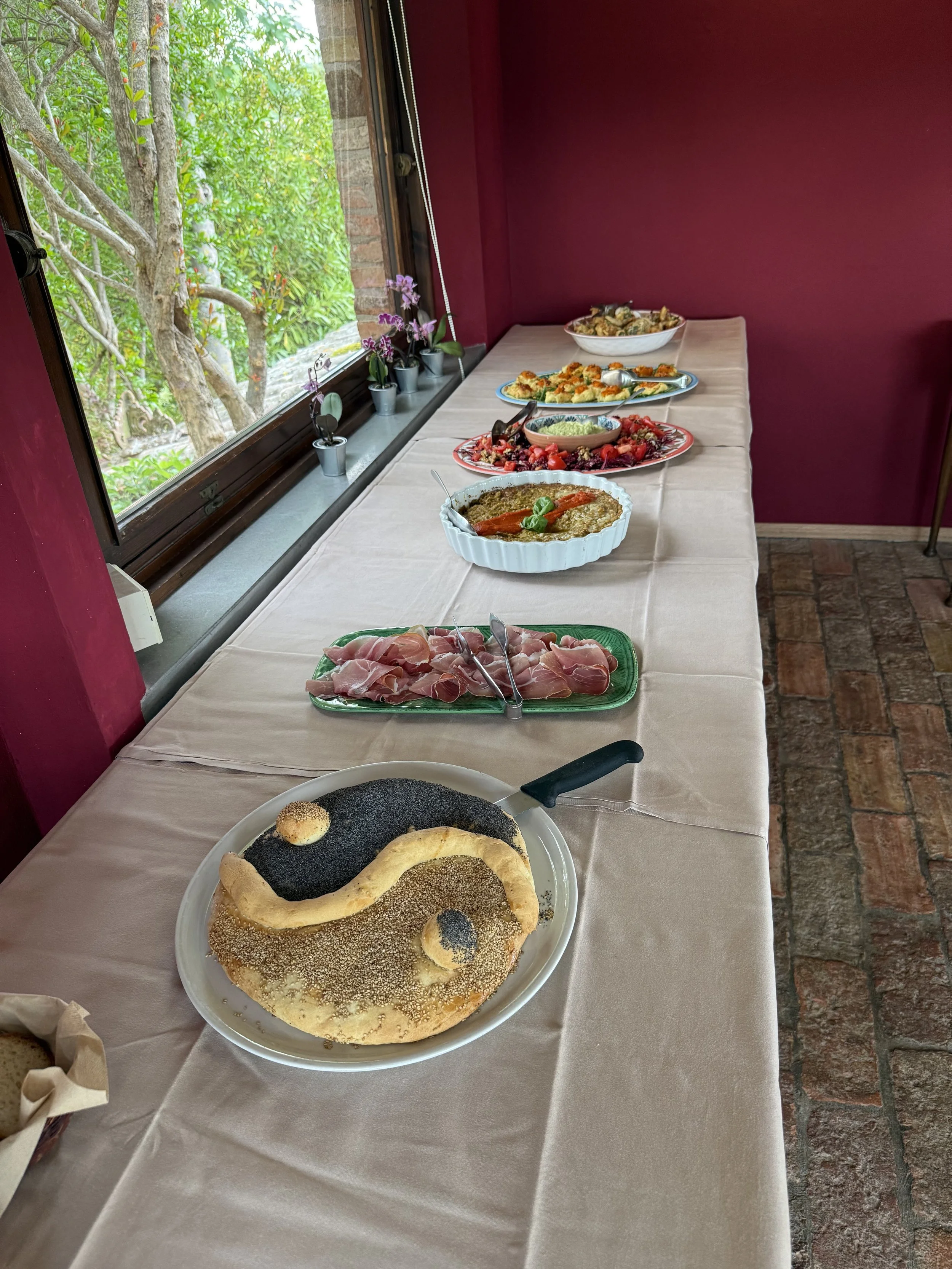 A table with various breakfast foods including a large flatbread with sesame and poppy seeds, sliced meats, colorful salads, and baked dishes, set indoors near a window with potted plants and a view of trees outside.