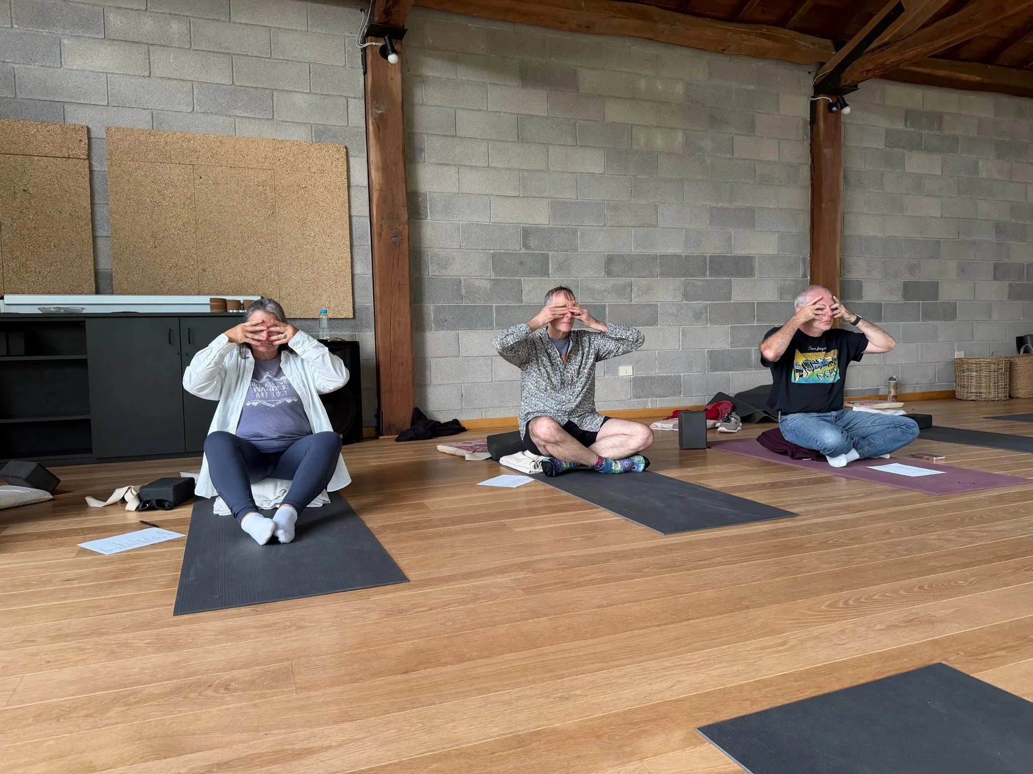 Three people practicing yoga in a studio, sitting cross-legged on mats with palms pressed together in front of faces, in a meditation pose.