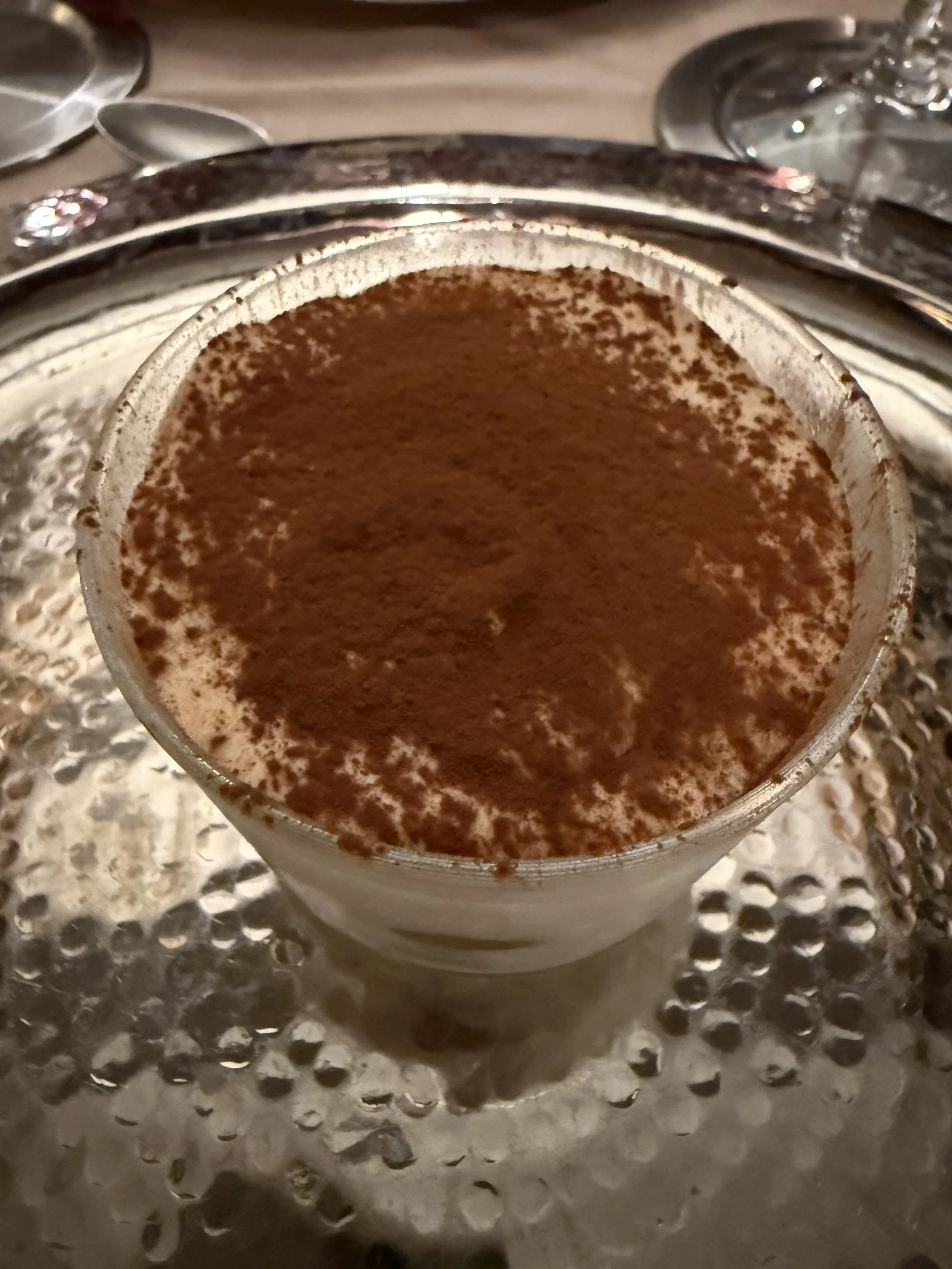 A close-up of a glass cup of hot coffee topped with cocoa powder, placed on a textured metallic surface with spoons visible in the background.