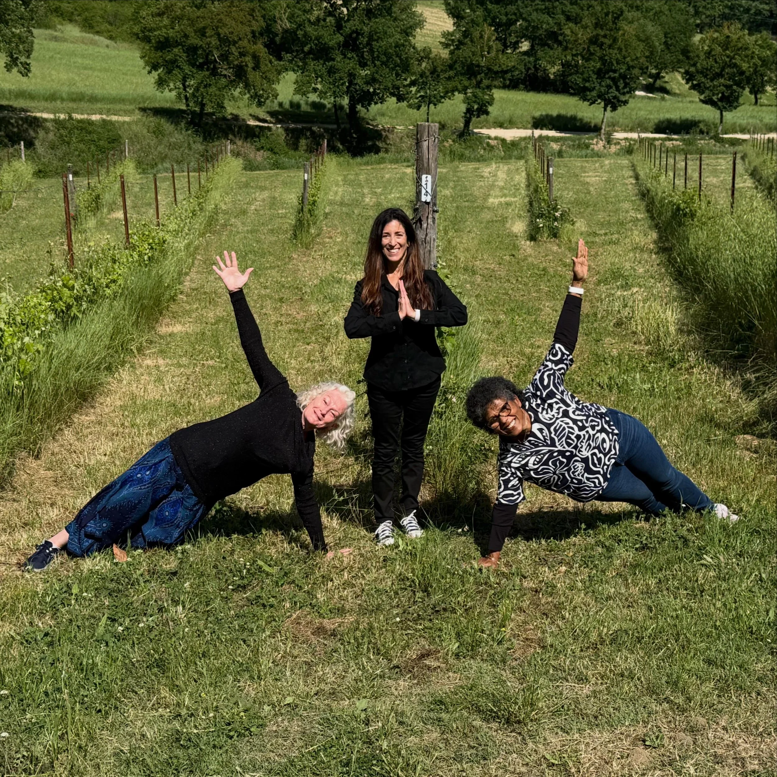 A group of people practicing yoga outdoors in a grassy field with trees and hills in the background. One person in the center is smiling and has hands in a prayer position, with others around her in different yoga poses.