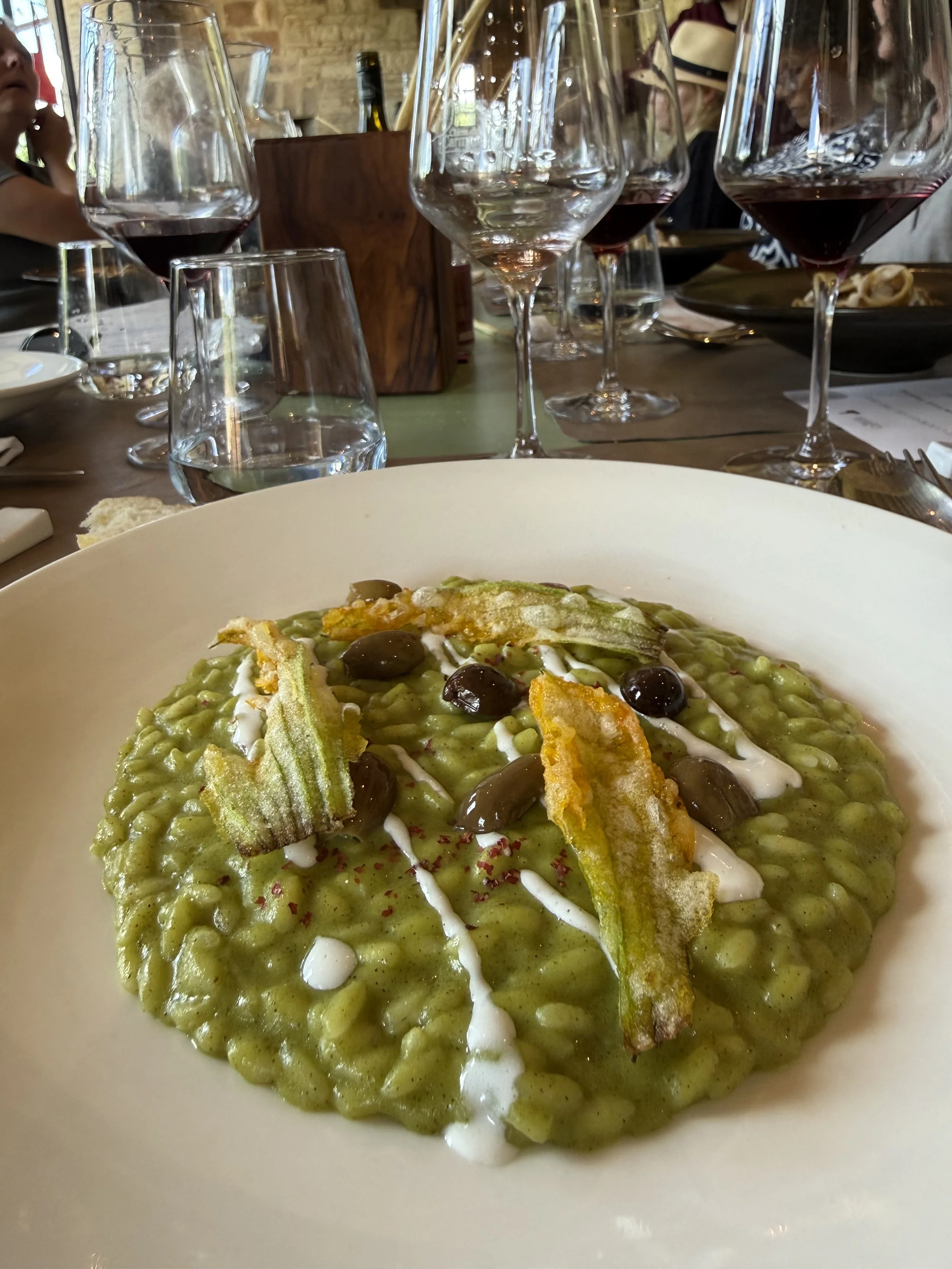 A bowl of green risotto topped with fried zucchini chips, black olives, and drizzled with white sauce, placed on a restaurant table with wine glasses and bottles in the background.