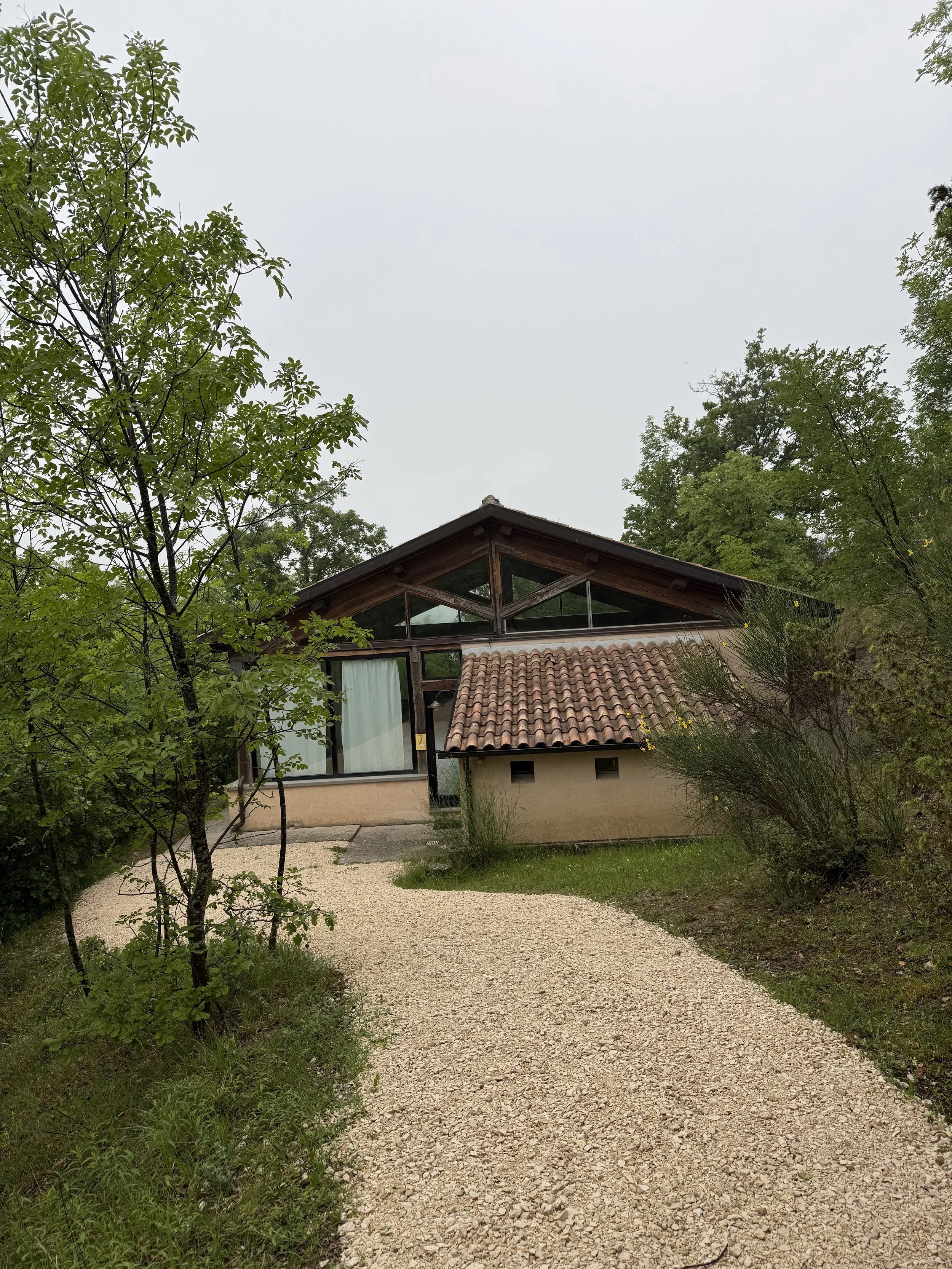 A house with a sloped roof and large windows is seen behind a gravel path, surrounded by green trees and bushes, with overcast sky.