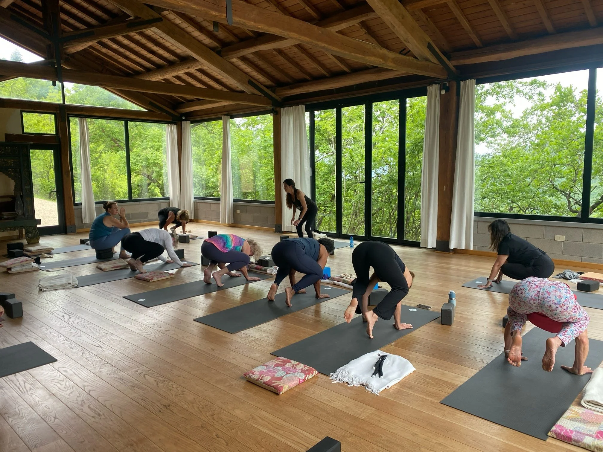 A group of people participating in a yoga or meditation class inside a spacious room with large windows showing green trees outside. They are in various poses on mats and cushions.
