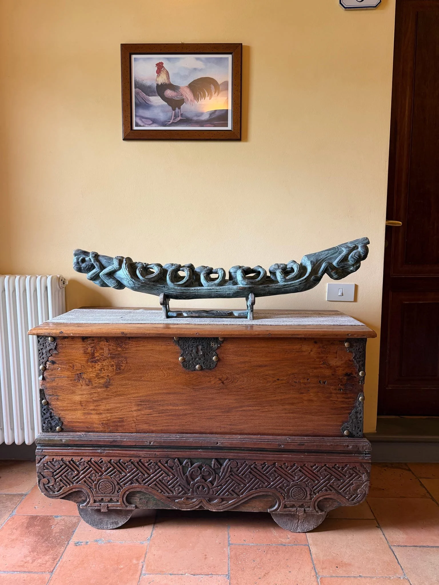 A wooden antique chest with carved decorative details on the front and sides, topped with a carved wooden bowl. Above the chest, on the yellow wall, is a framed painting of a rooster standing outdoors at sunset."