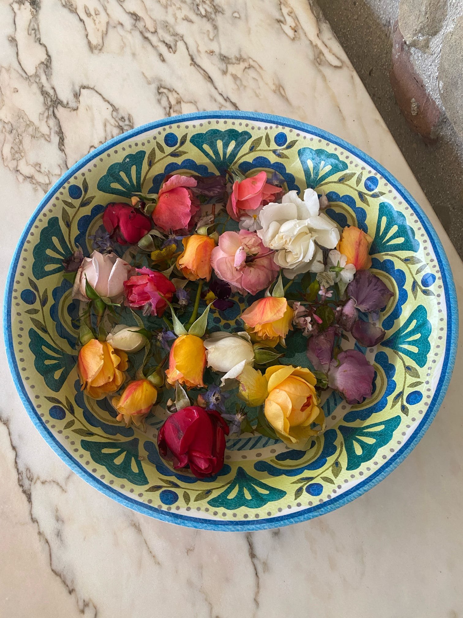 A decorative bowl with a blue and yellow pattern filled with multicolored rose petals, placed on a marble surface.