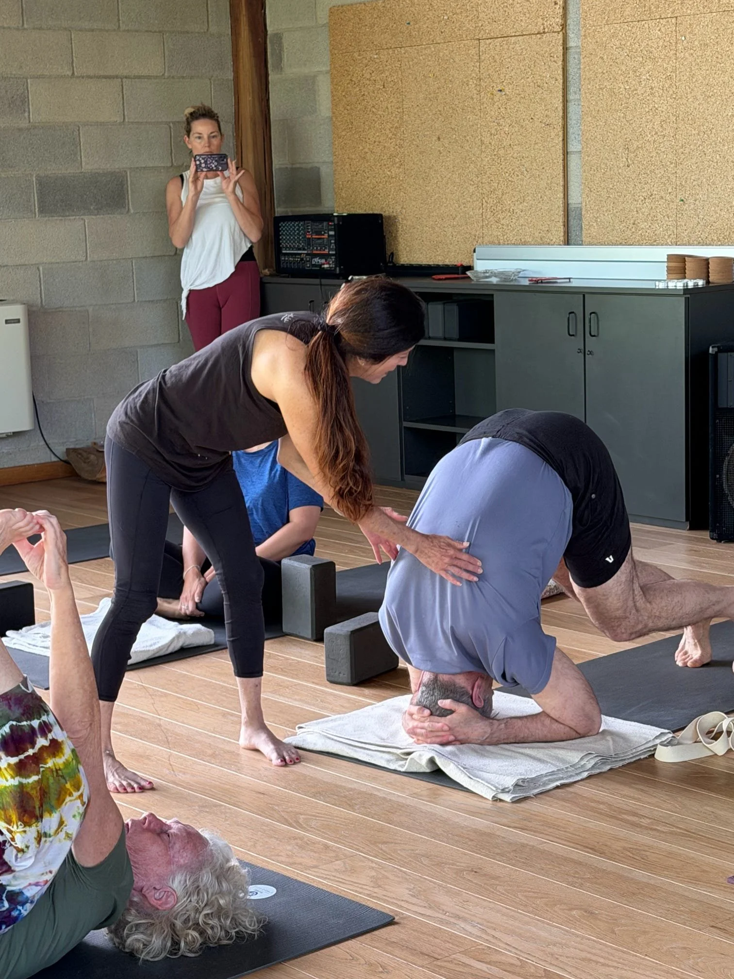 People participating in a yoga class focusing on flexibility and balance exercises, with some assisting others and a woman taking photos in the background.