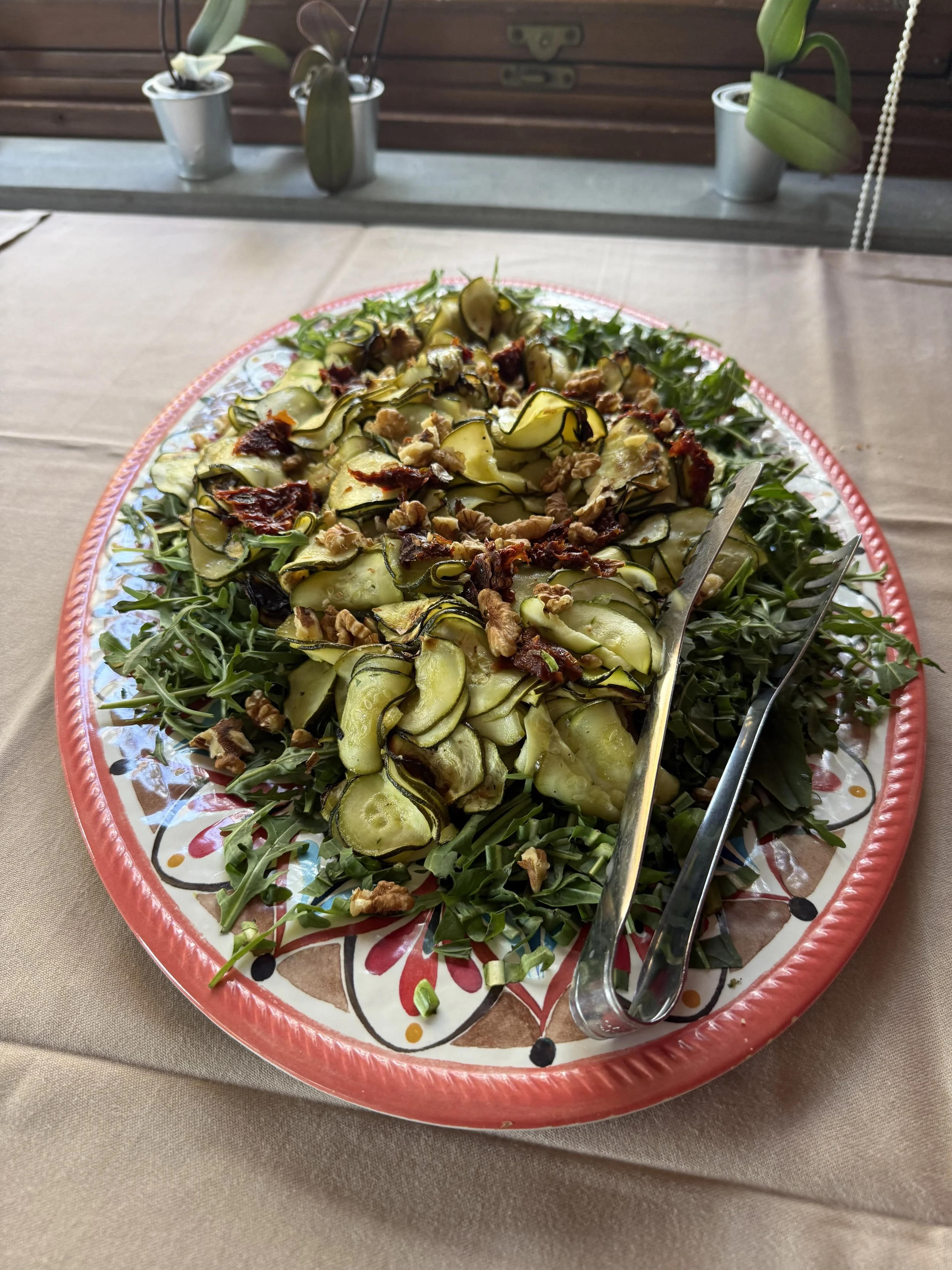 A colorful platter of sliced zucchini and other vegetables, garnished with nuts and herbs, on a decorative plate on a beige tablecloth, with plants in pots in the background.