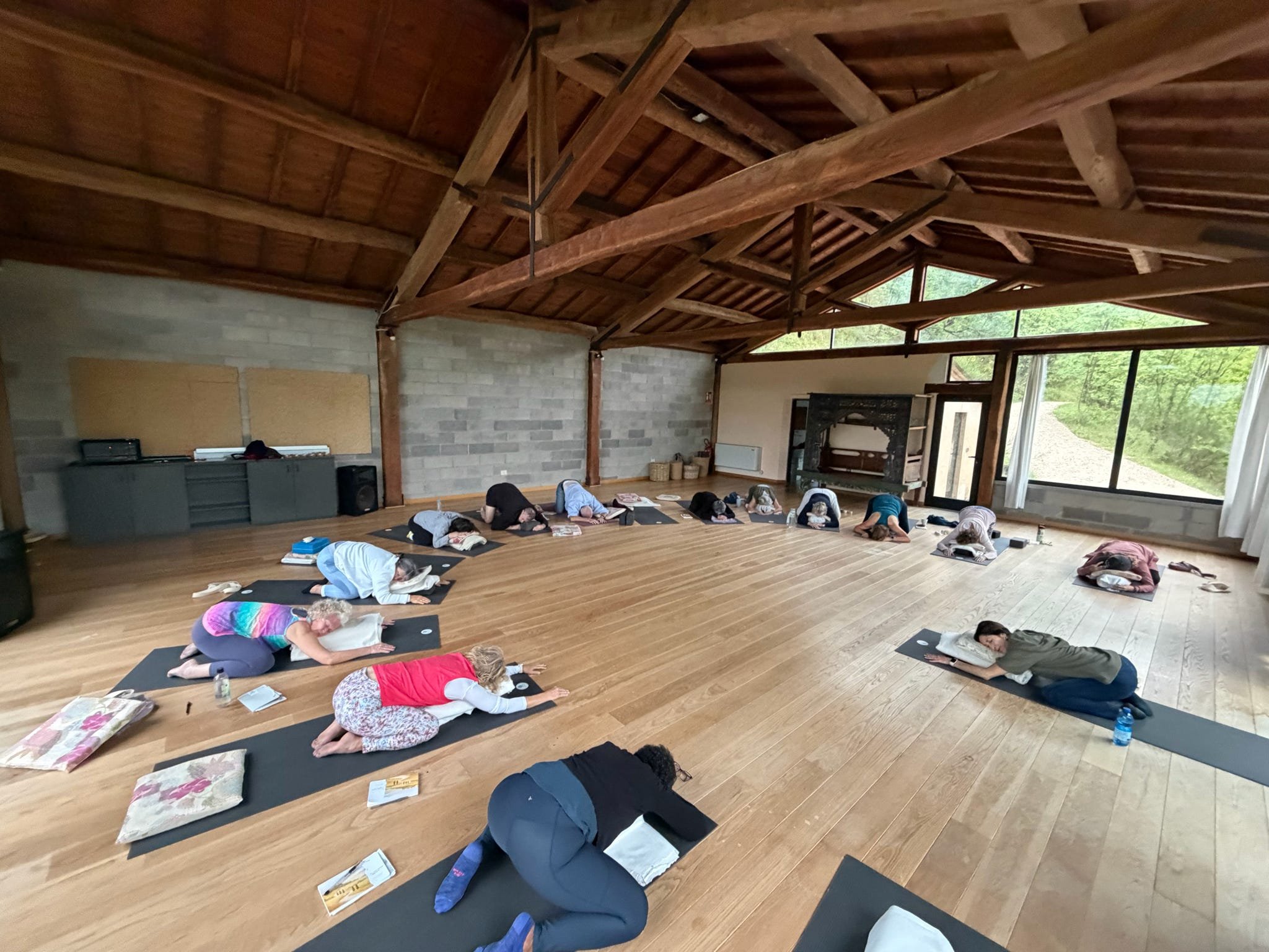 A group of people participating in a yoga class in a spacious room with large windows, wooden ceiling, and hardwood floors, all in a kneeling yoga pose on black mats.