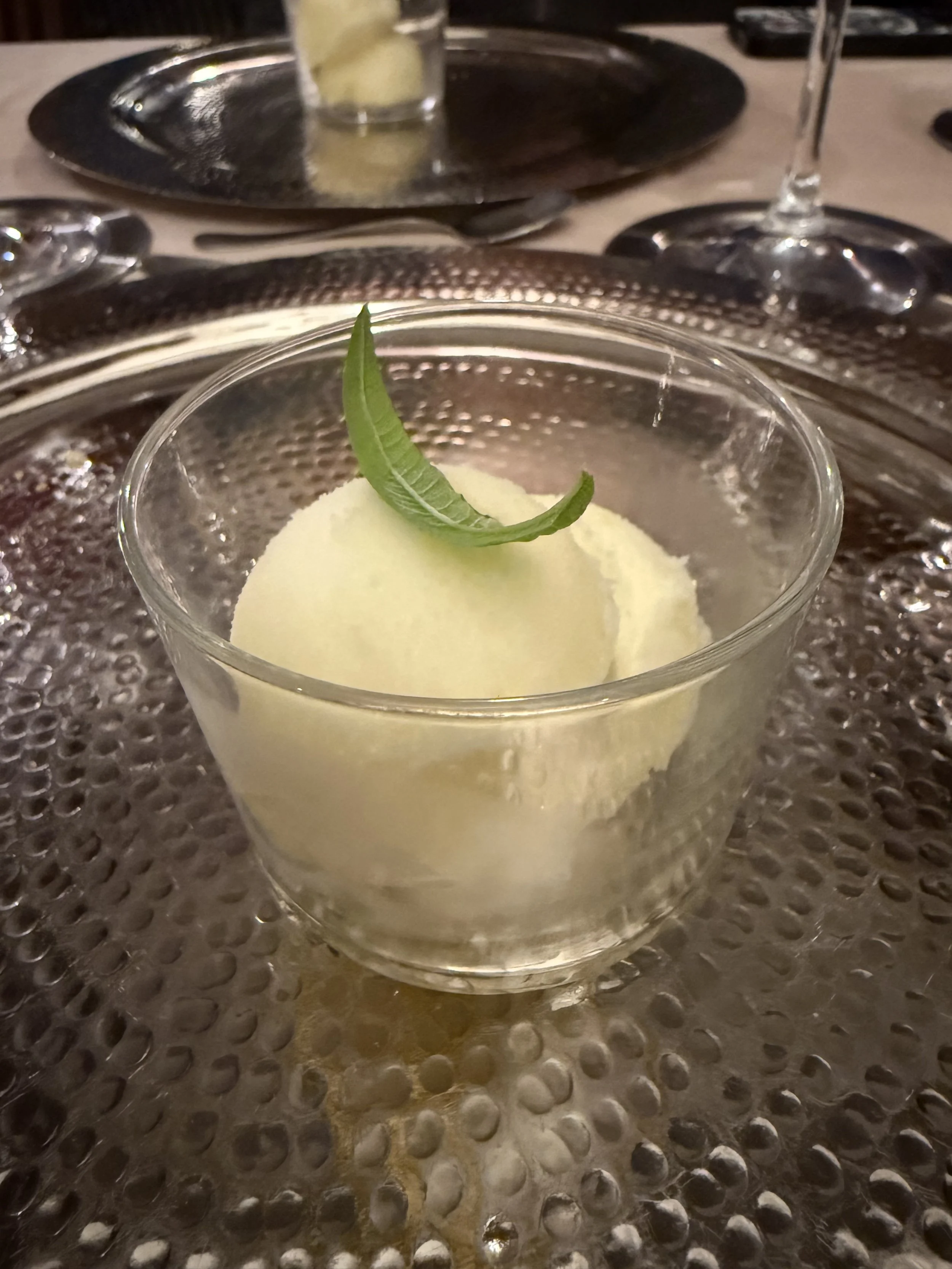 A glass of yellow frozen dessert garnished with a small green leaf, placed on a textured metal table or countertop.