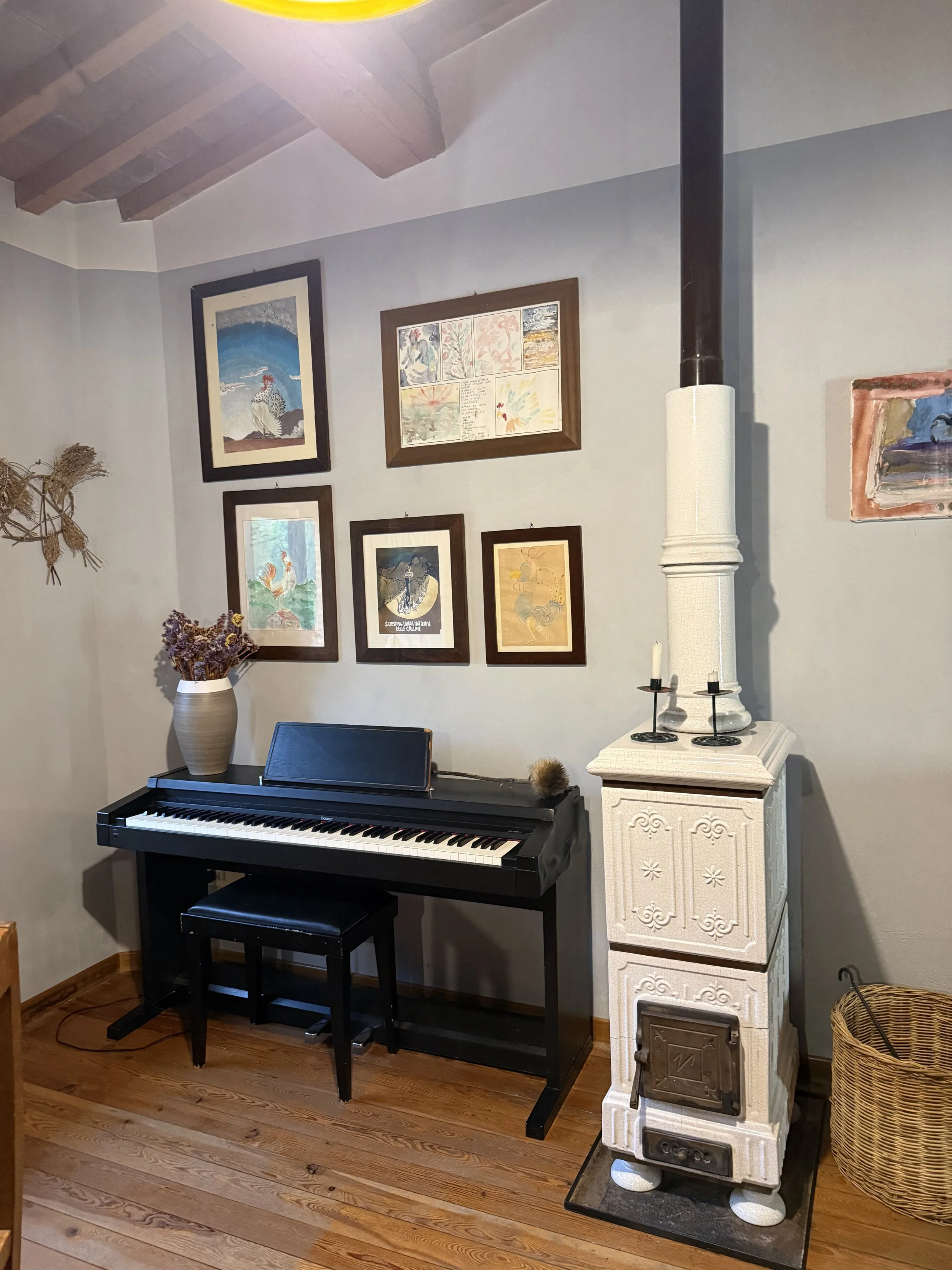 Interior of a room with a black digital piano against a light gray wall. Above the piano, there are several framed pictures and artwork. To the right of the piano, there is a white decorative old-fashioned stove and a woven basket. To the left, a lar